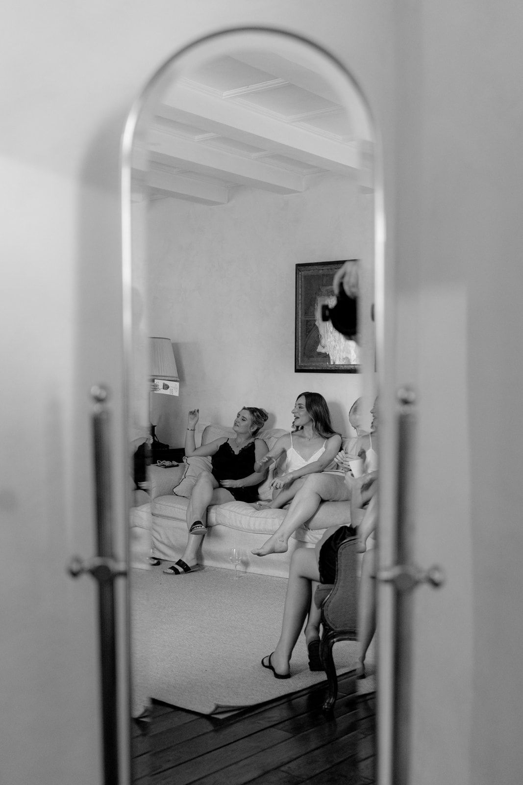 Black and white mirror reflection of women laughing on a couch during bridal party preparations.