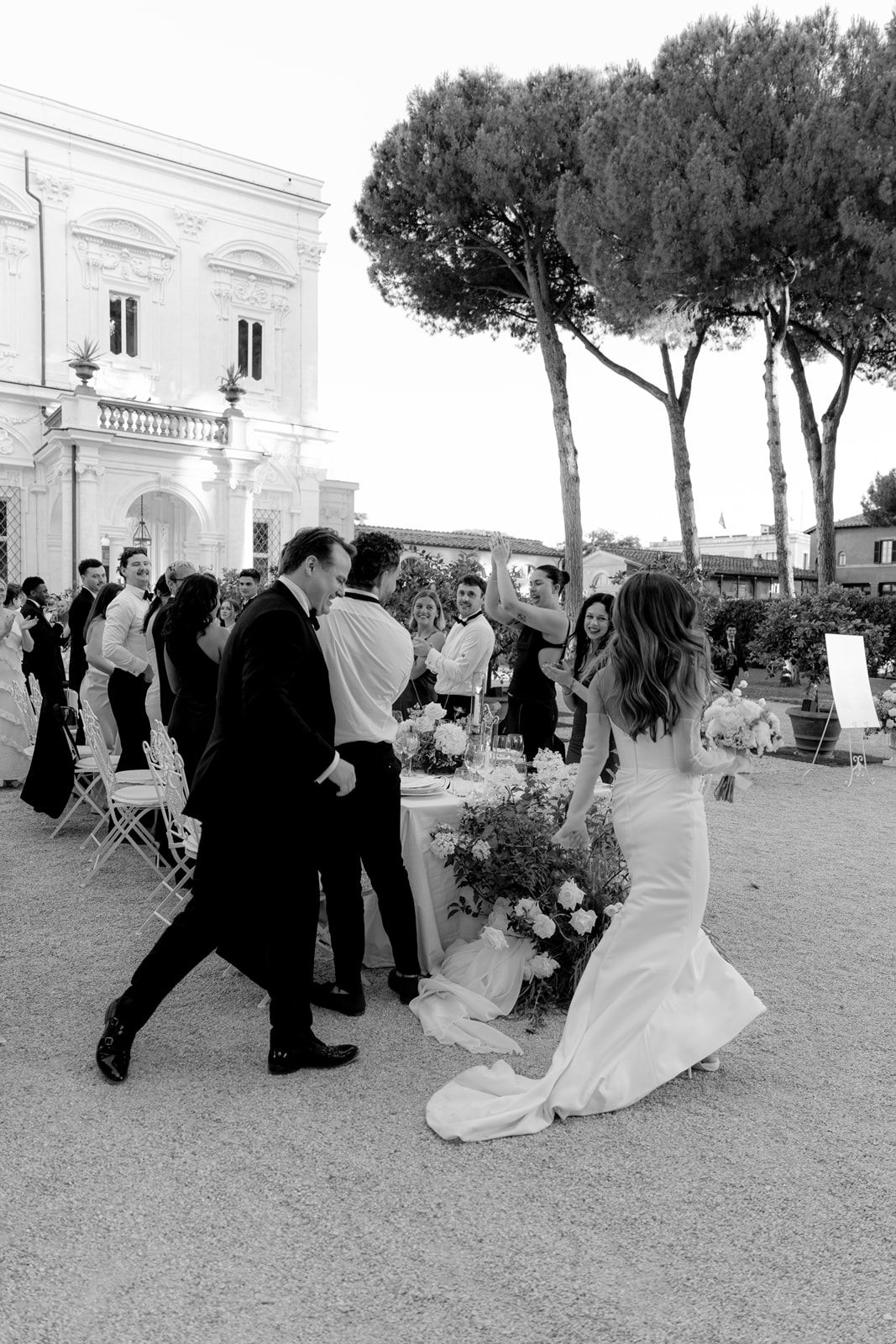 Black and white photo of a bride in a fitted gown greeting wedding guests outdoors at an elegant Italian villa.