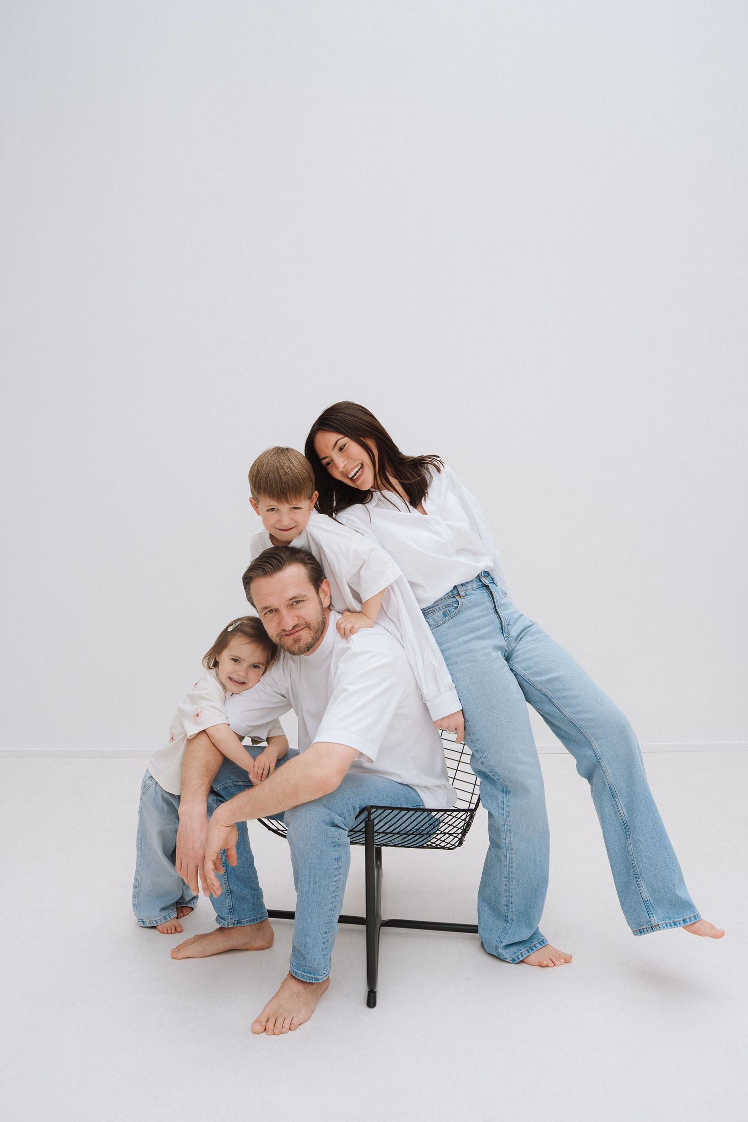 Happy family in white shirts and jeans posing together on a chair against a white background.