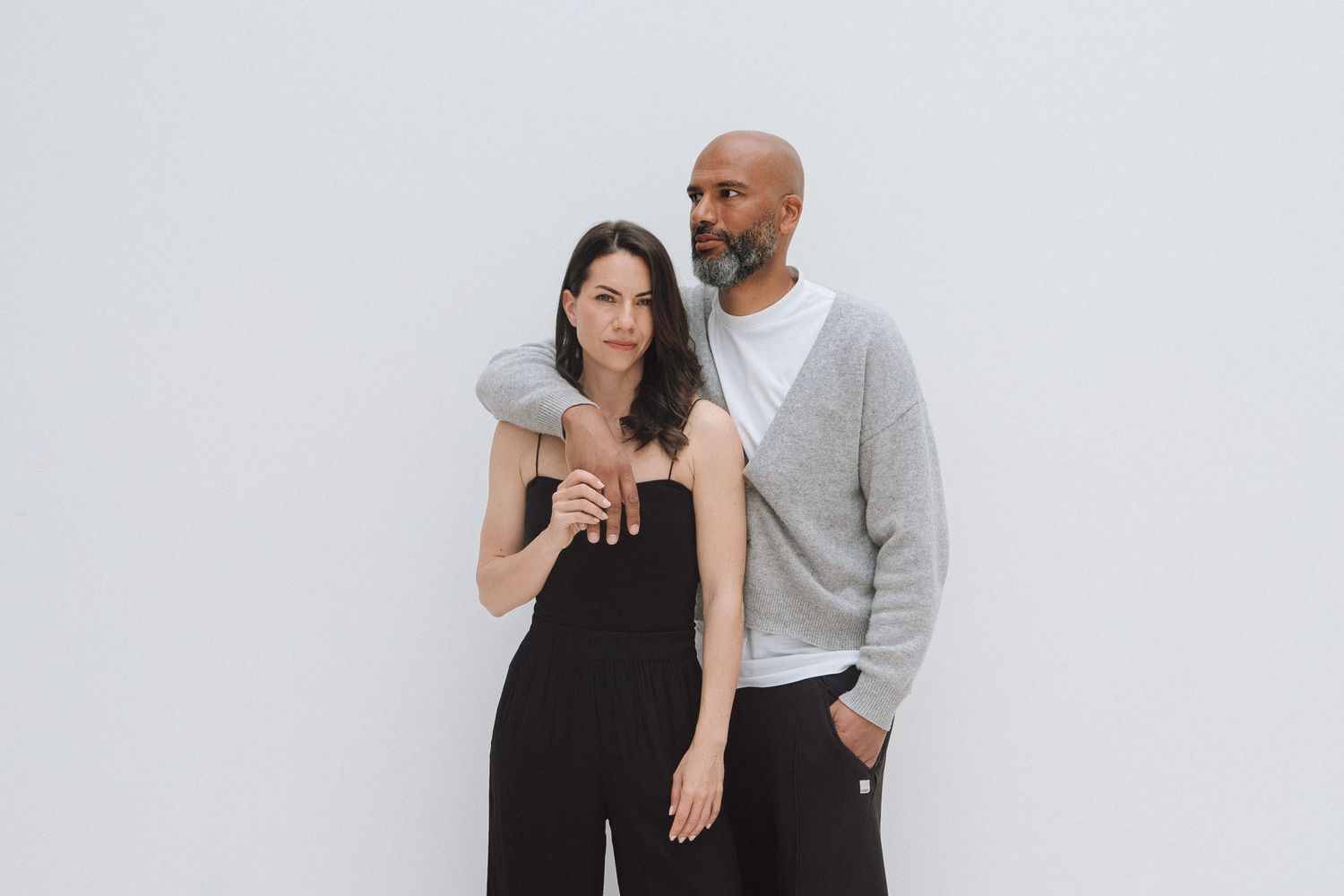 A man and woman pose together against a white background, she in a black jumpsuit, he in a grey sweater.