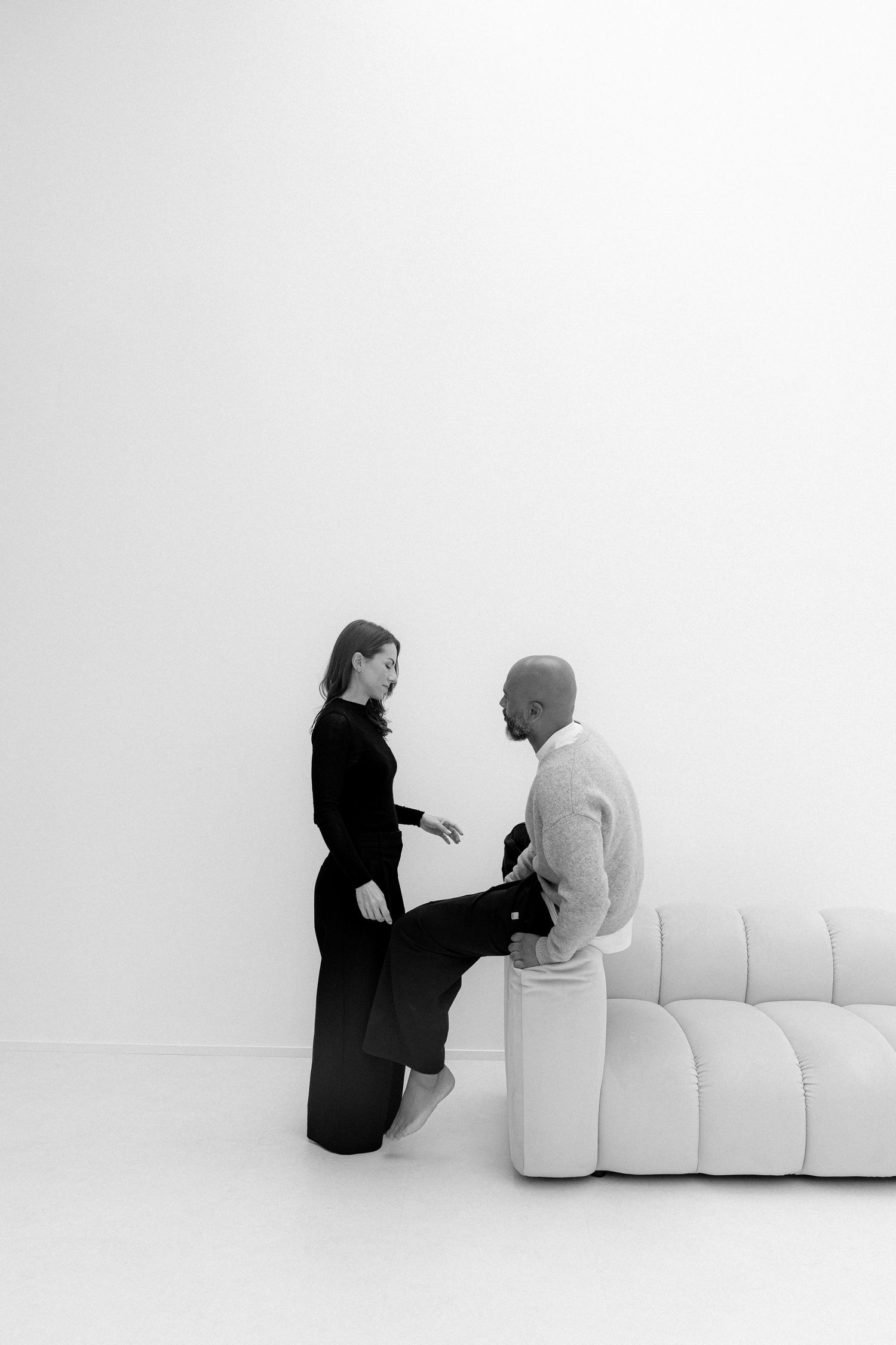 Black and white photo of a man and woman in conversation beside a white curved sofa in a minimalist room.