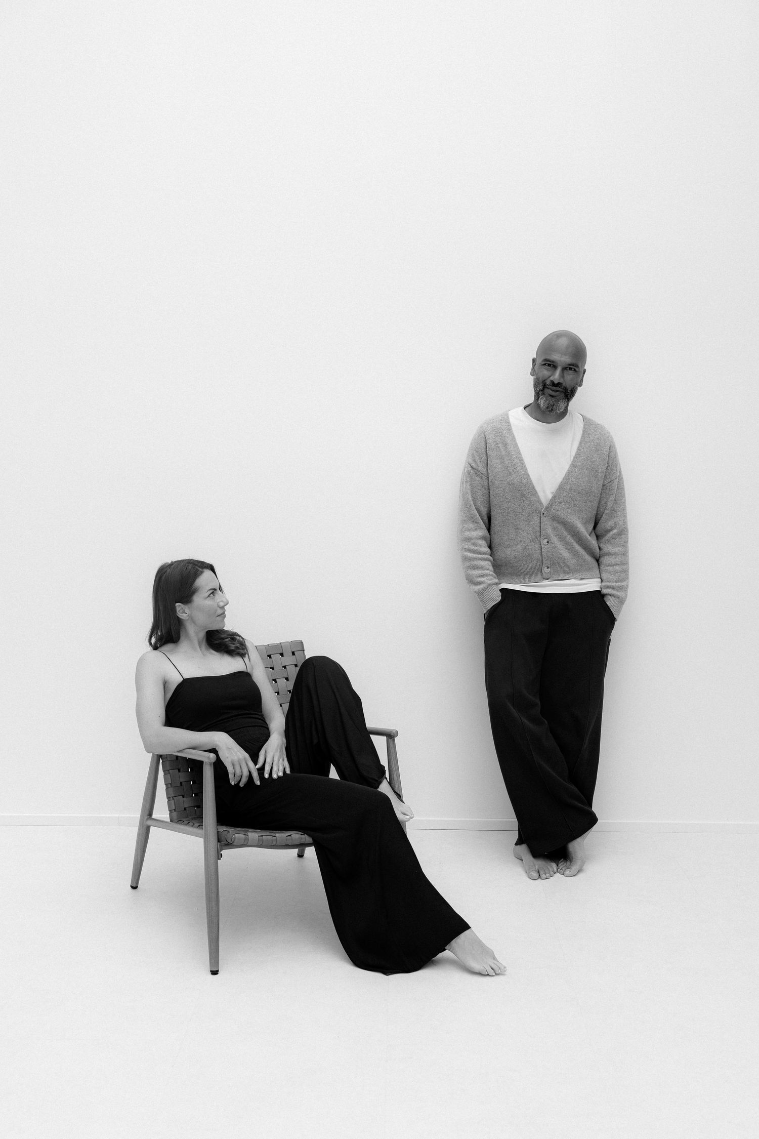 Black and white photo of a woman sitting in a chair and a bald man leaning against a white wall.