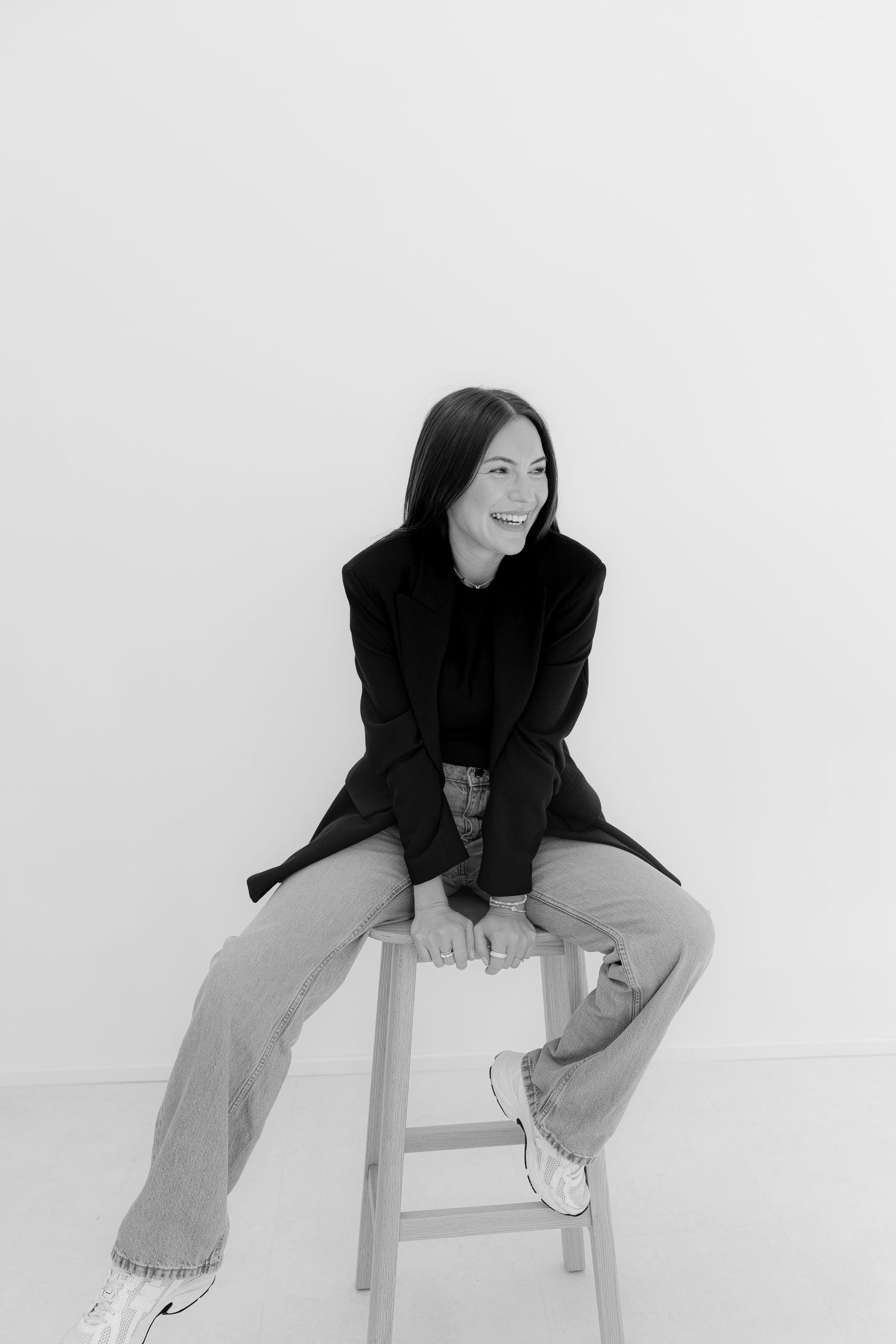 Woman in black blazer and wide-leg pants sits smiling on a stool against a white background.
