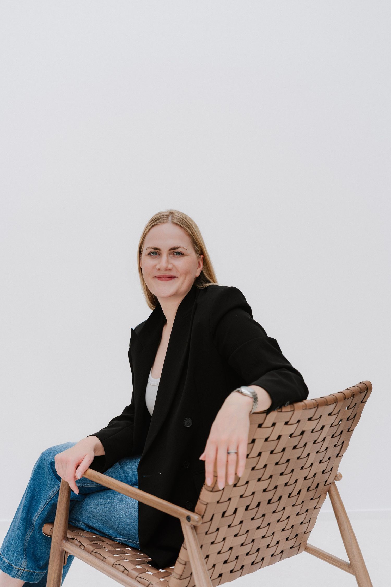 Woman in black blazer and jeans sitting relaxed in a woven wooden chair against a white background.