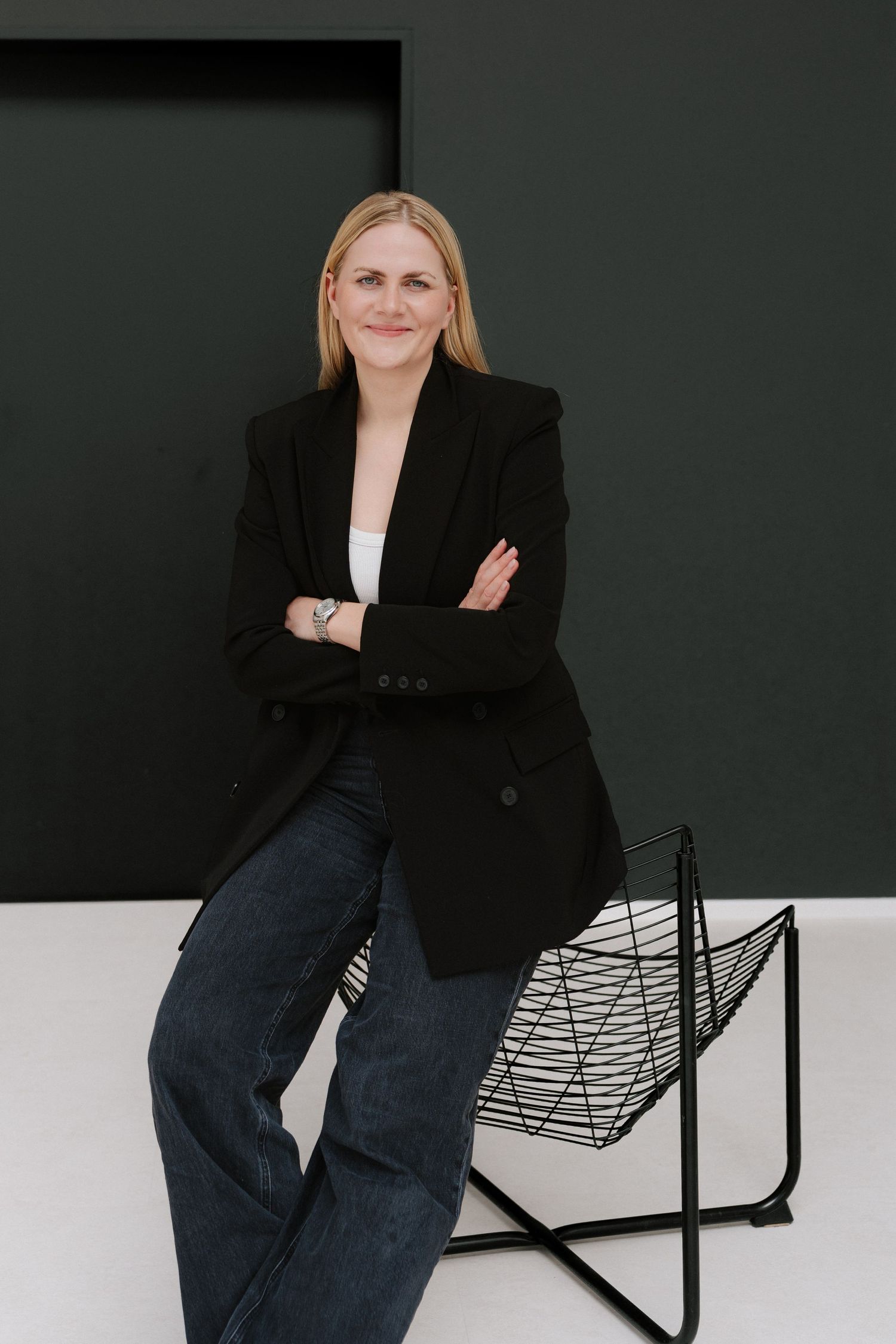 Blonde woman in black blazer and jeans poses with arms crossed beside a wire chair against dark wall.