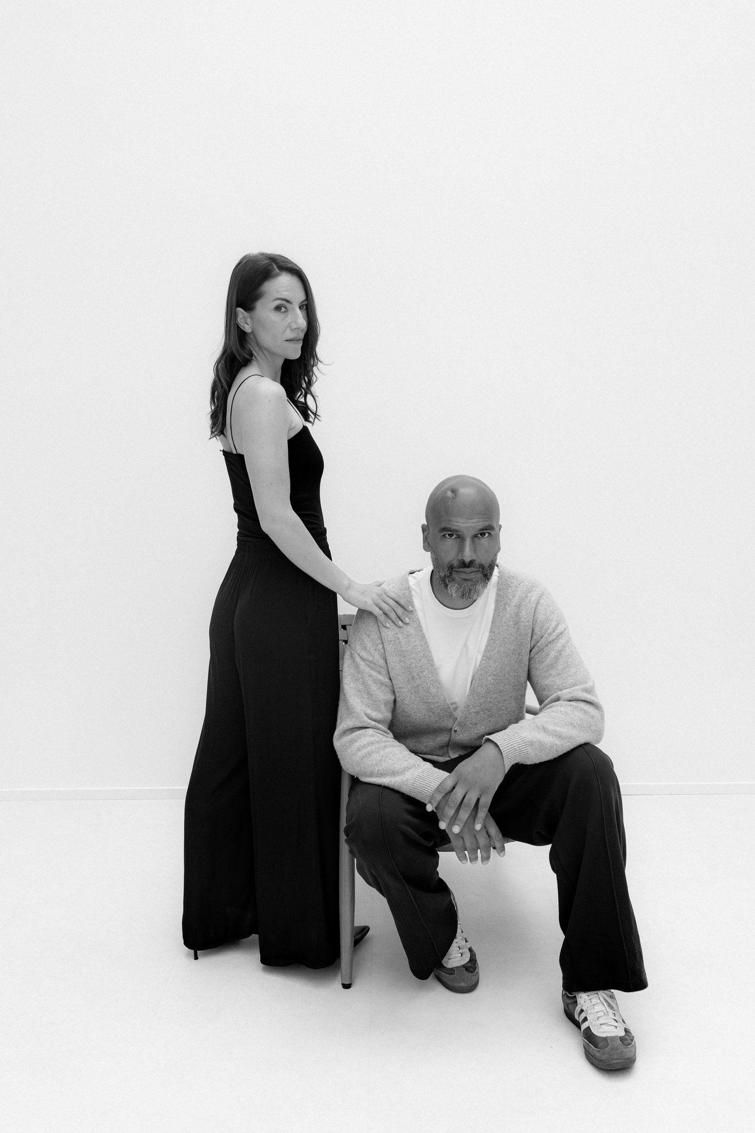 Black and white studio portrait of a woman in black jumpsuit standing beside a seated bald man in cardigan.