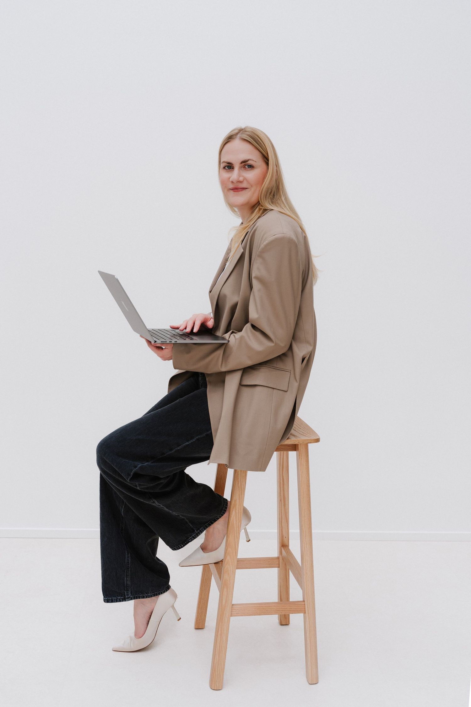 Professional blonde woman in beige blazer sitting on wooden stool with laptop, smiling against white background.