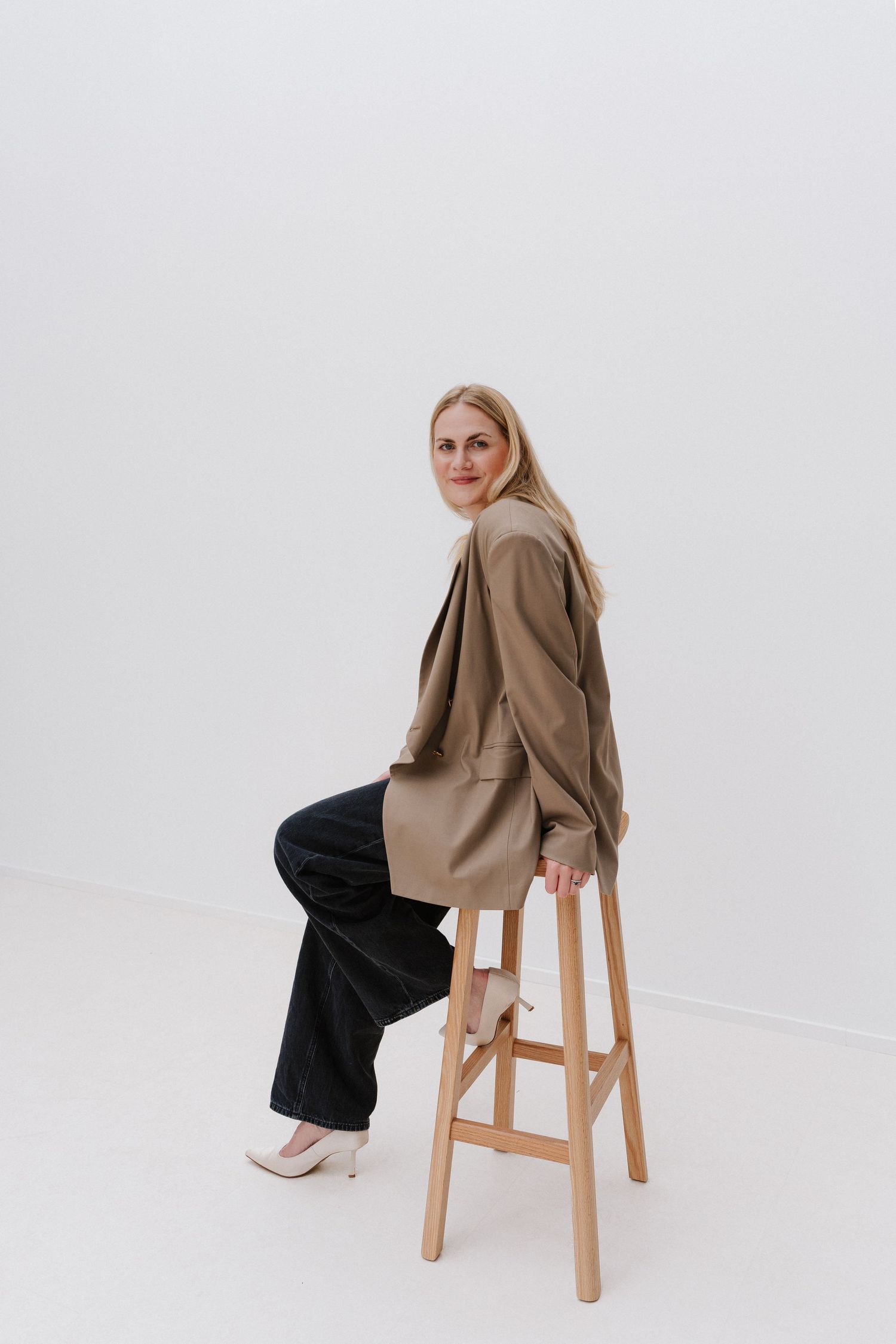 Blonde woman in beige blazer and black pants sitting on wooden stool, smiling over shoulder against white background.