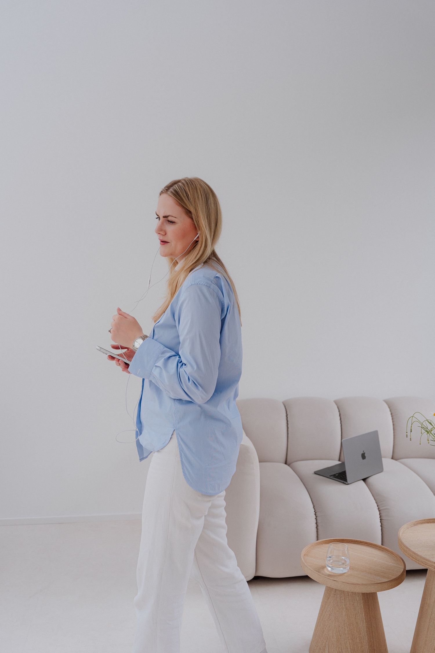 Blonde woman in blue blazer and white pants holding a notebook, standing near a beige sofa with a laptop.
