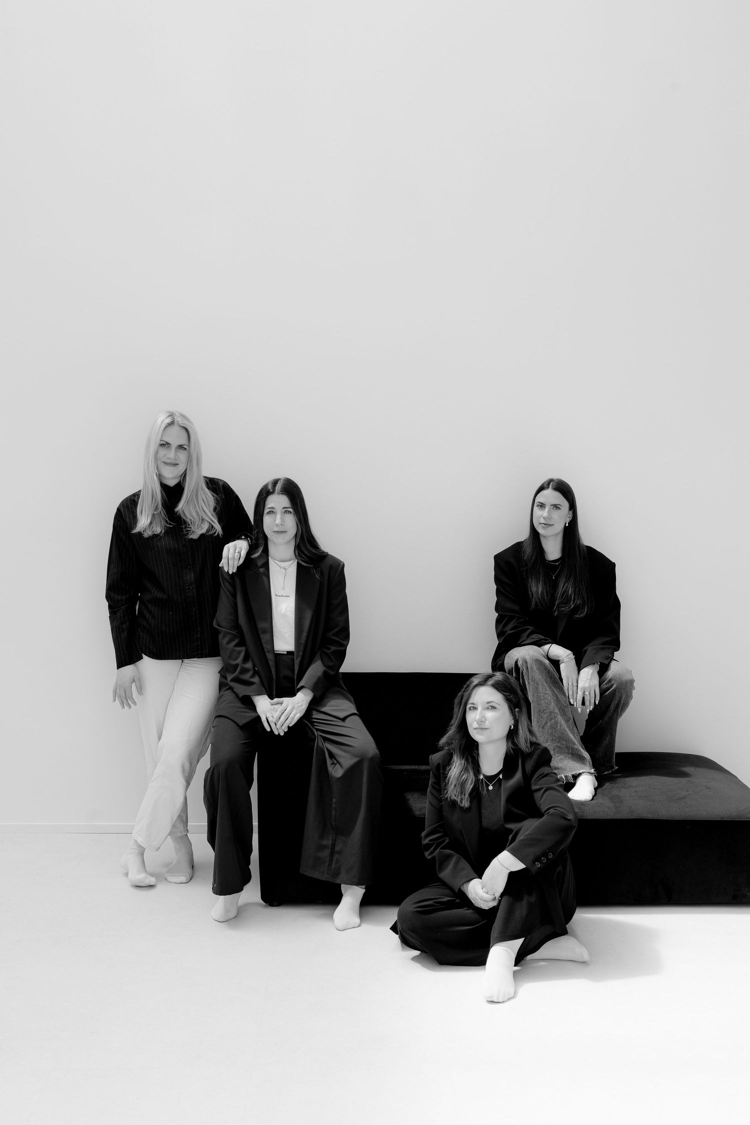 Four women in black outfits pose together in a minimalist studio setting in a black and white photo.
