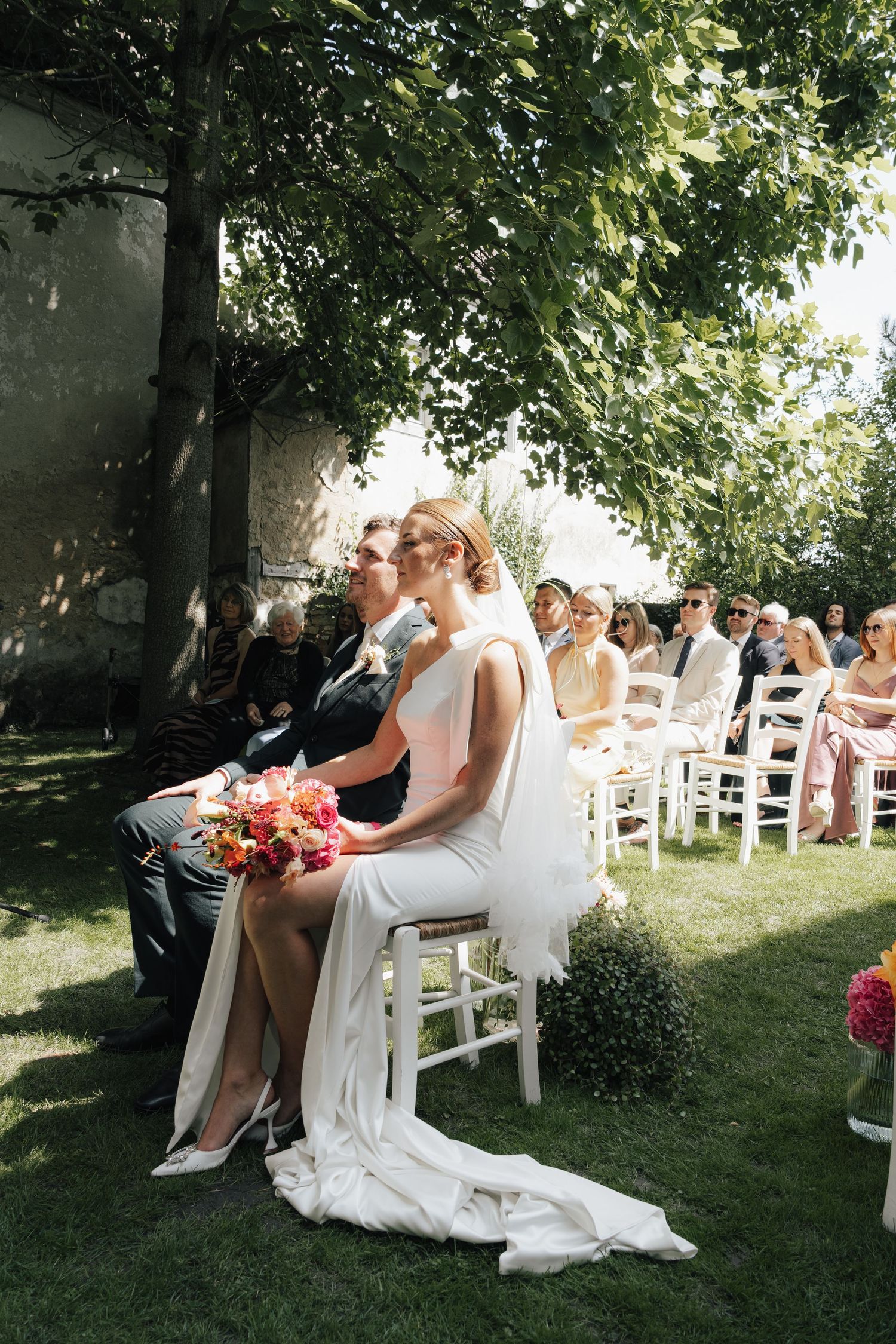 Wedding ceremony in a sunlit garden with white chairs and natural greenery.