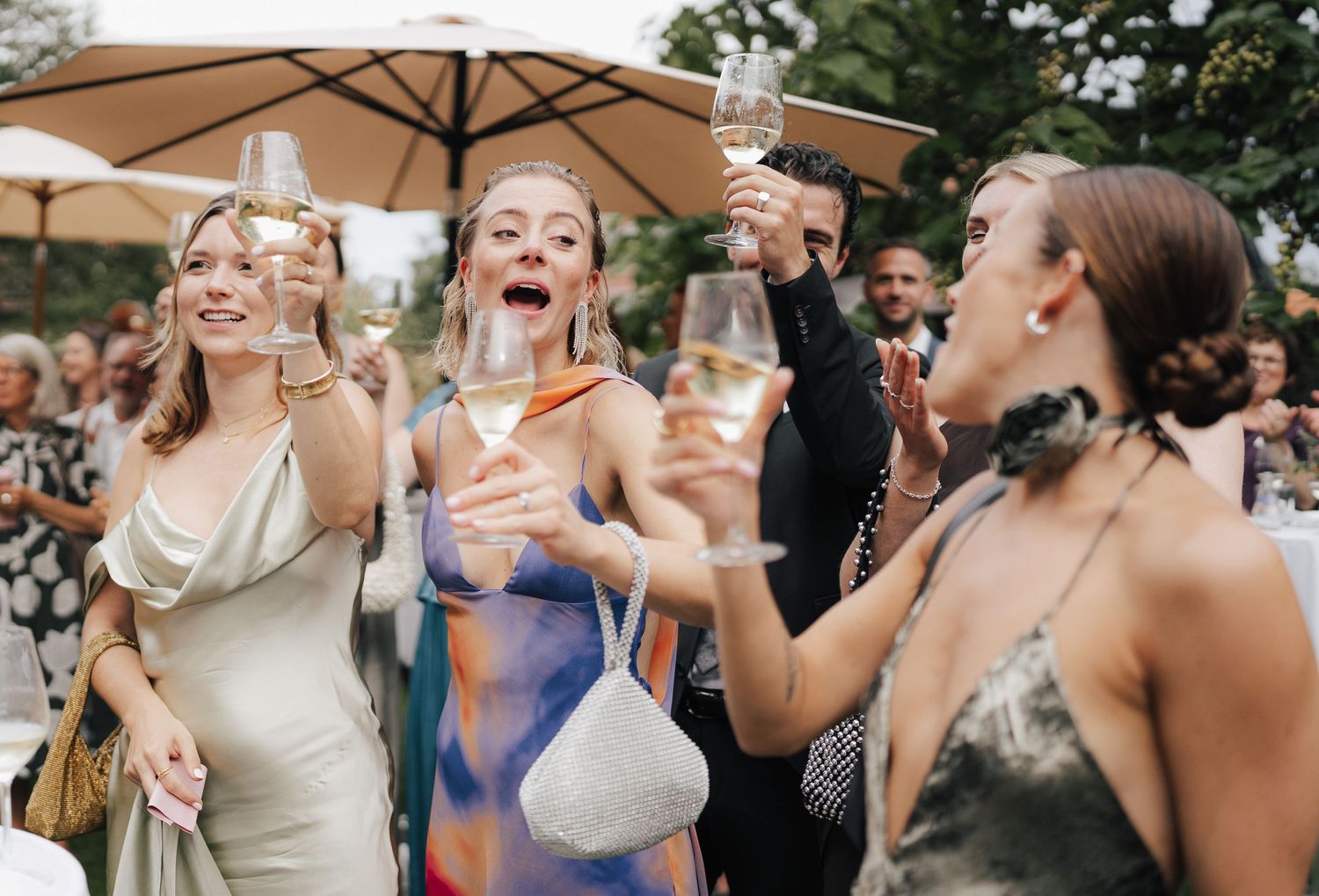 Wedding guests raise champagne glasses in celebration under a patio umbrella.