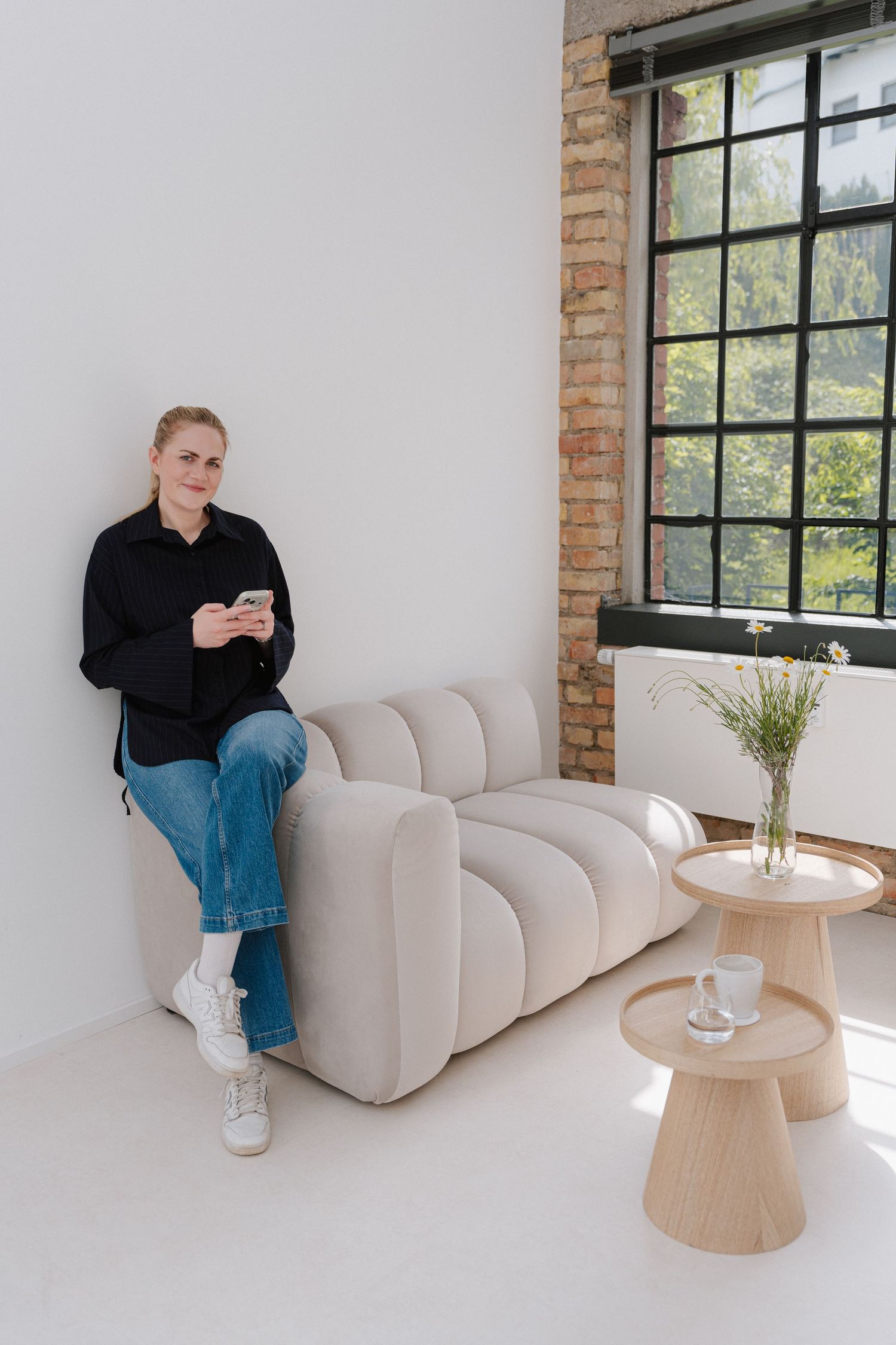 Woman in black shirt and jeans sitting on beige sofa using smartphone in modern white interior.