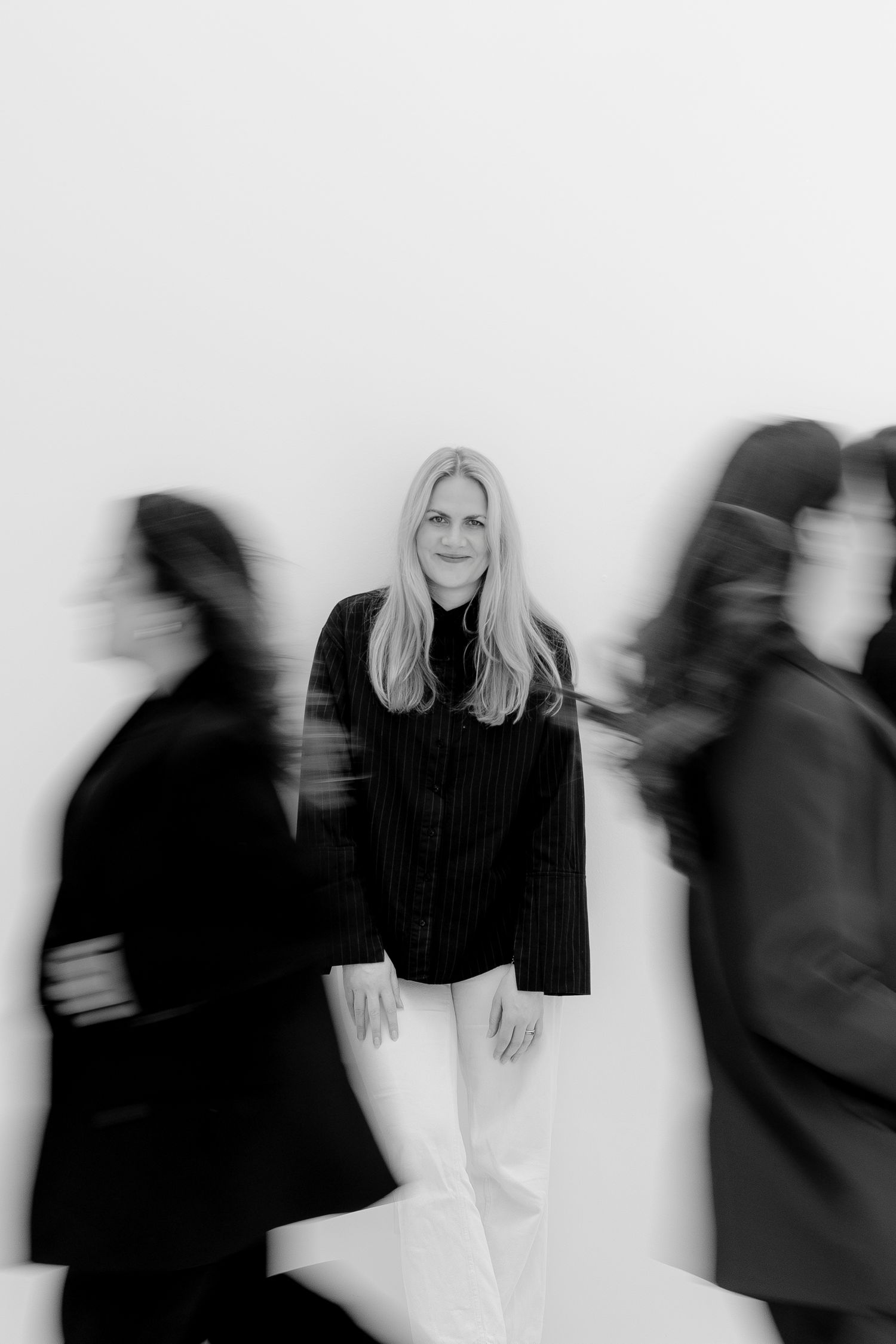 Black and white photo of blonde woman standing still among blurred moving figures in dark clothing.