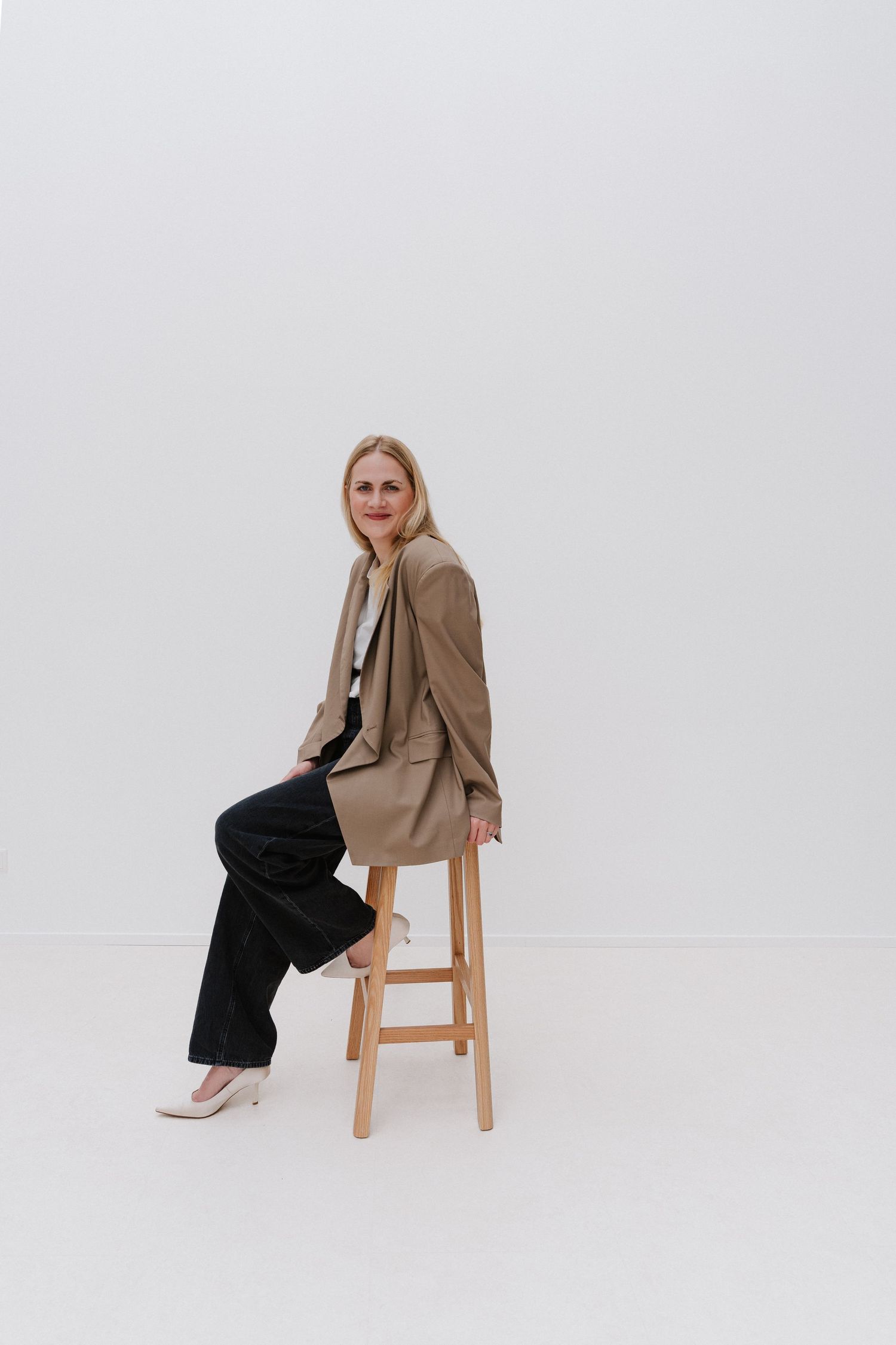 Blonde woman in beige blazer and black pants sitting on wooden stool against white background.