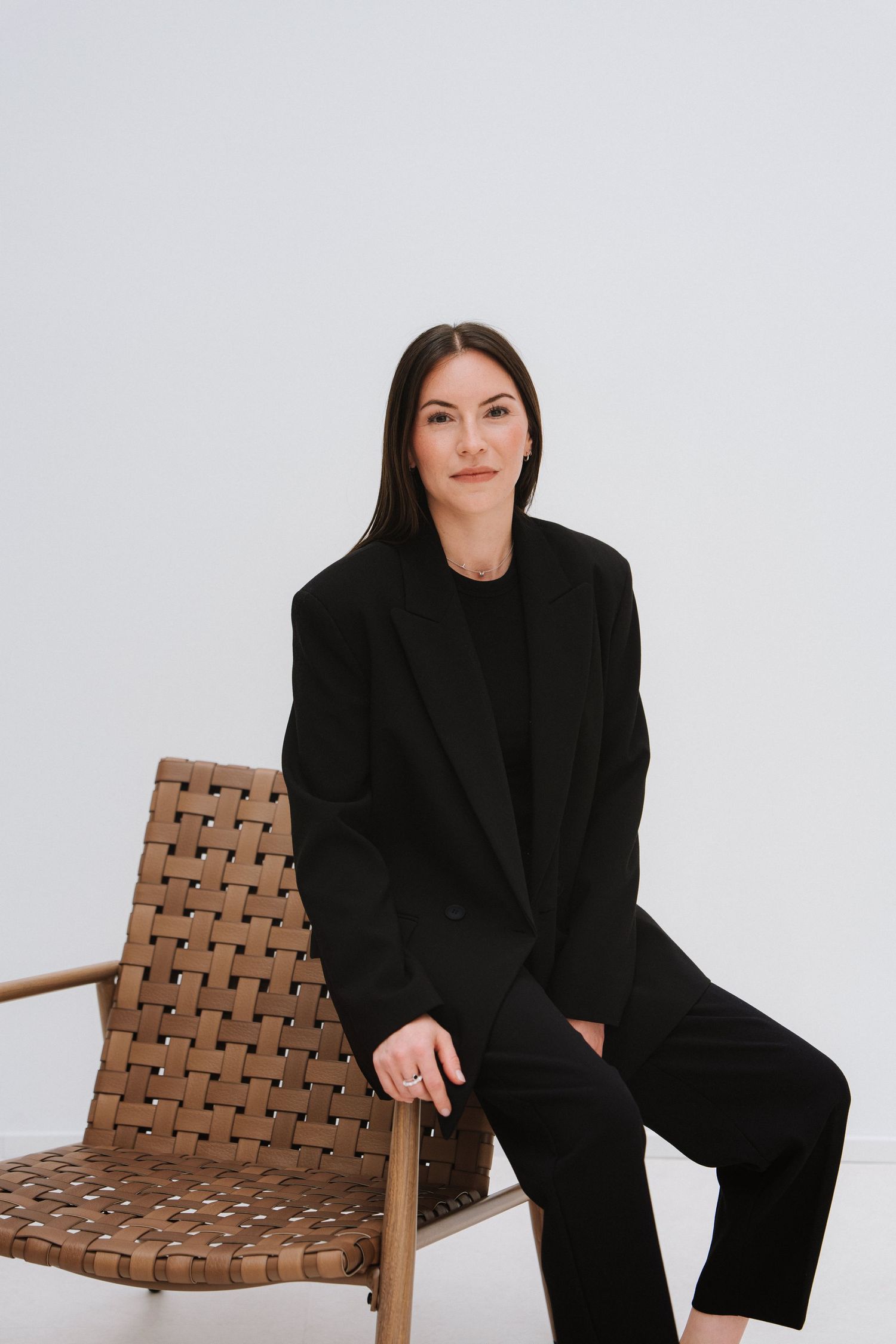 Woman in black suit sitting on woven leather chair against white background, professional portrait photo.