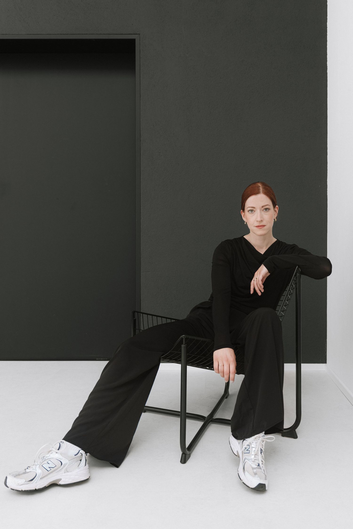 Woman in black outfit and white sneakers sitting on a dark metal chair against a gray wall.