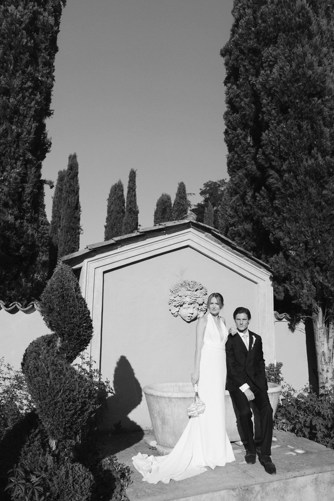 Black and white wedding photo of bride and groom posing by ornate garden wall with cypress trees.