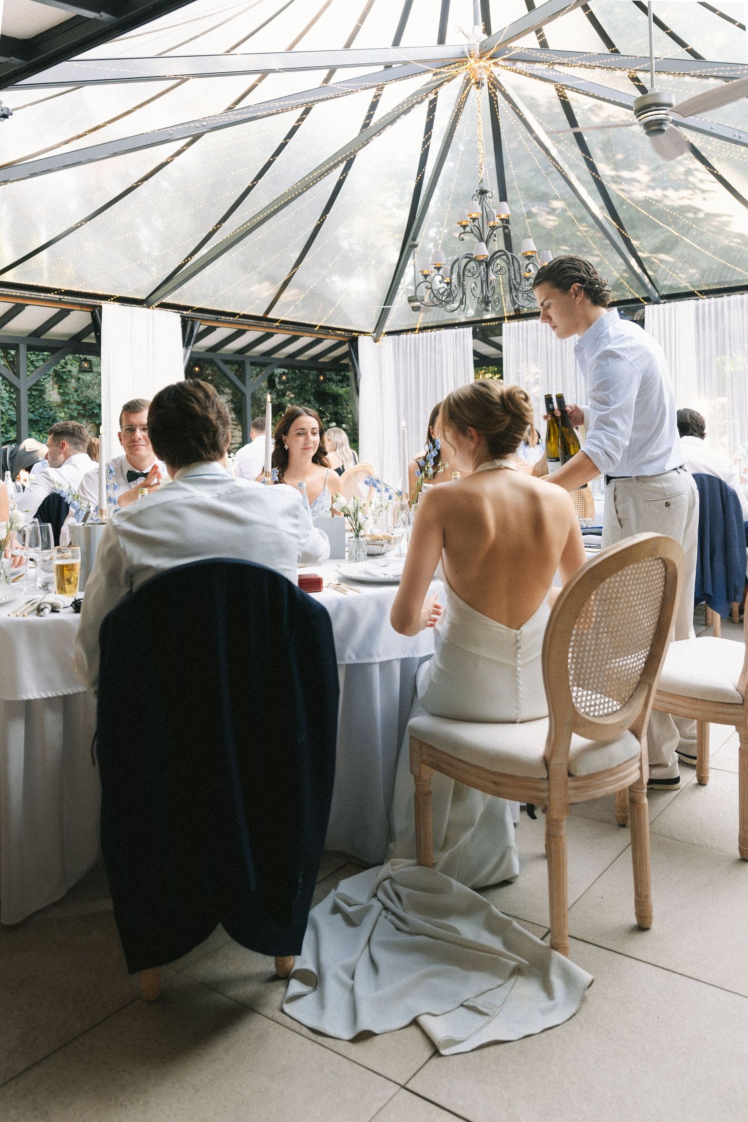 Bride and groom seated at elegant wedding reception table under glass ceiling, with waiter serving champagne.