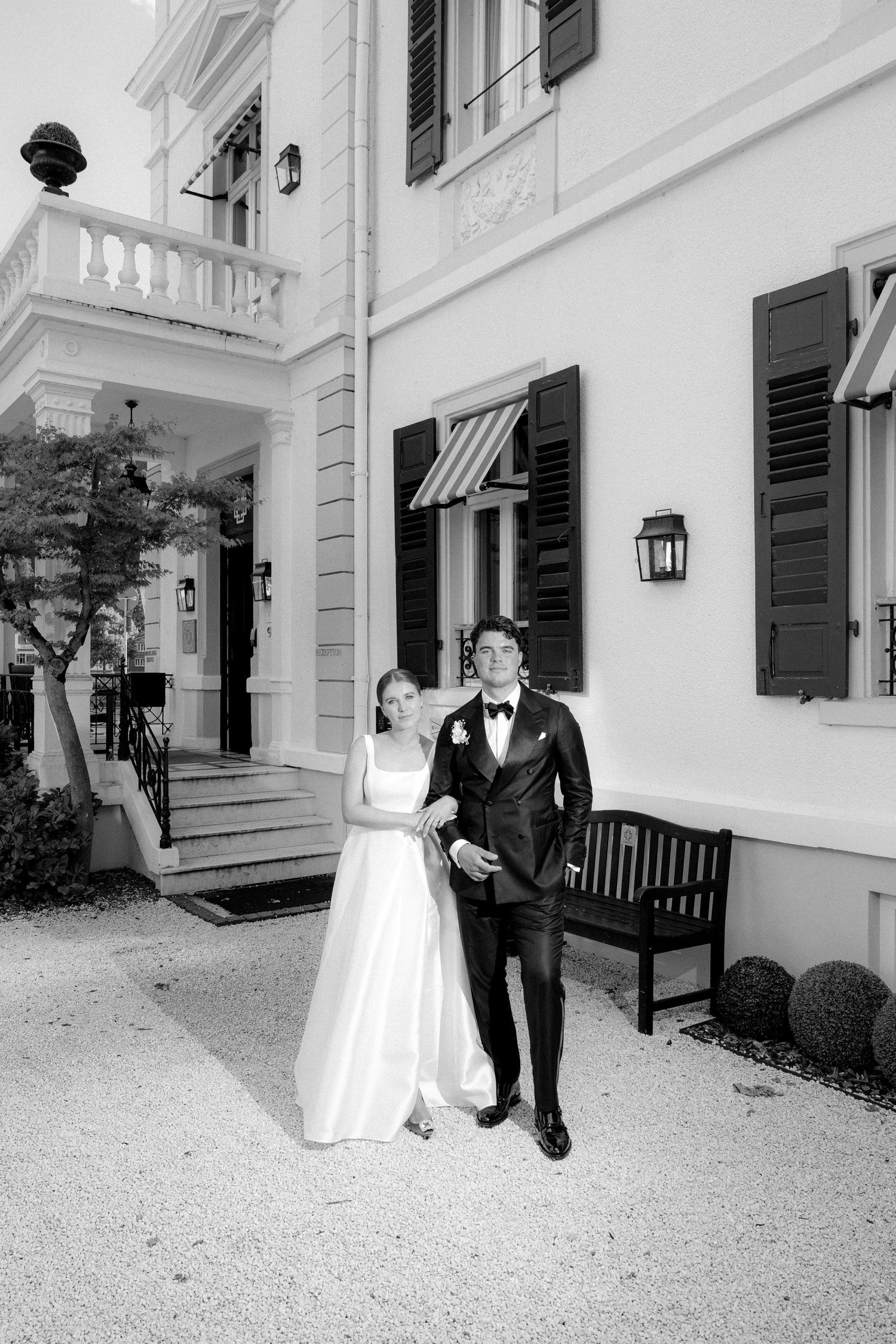 Wedding couple poses along white building exterior with black shutters and benches.