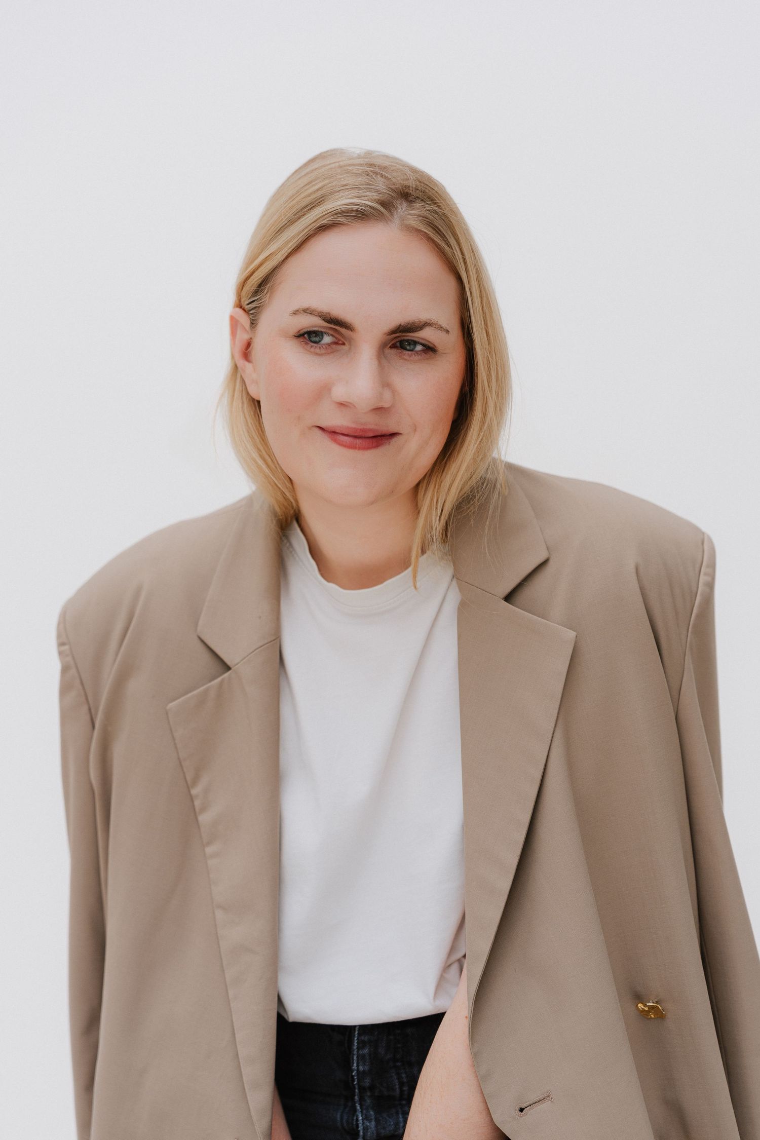 Blonde woman in beige oversized blazer and white t-shirt posing against light background.