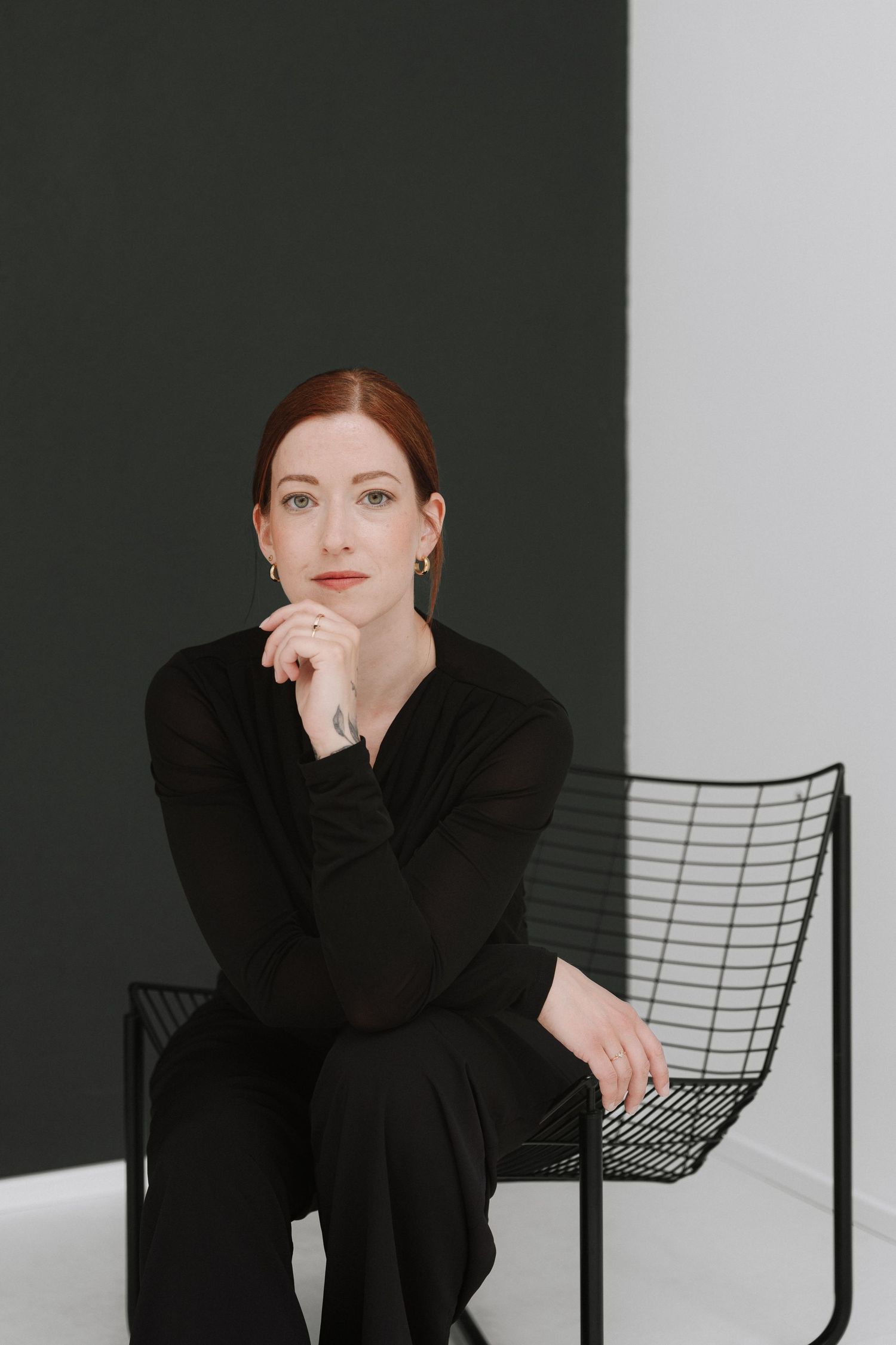 A woman with red hair in black outfit sits thoughtfully on a wire chair against a two-toned green and white wall.