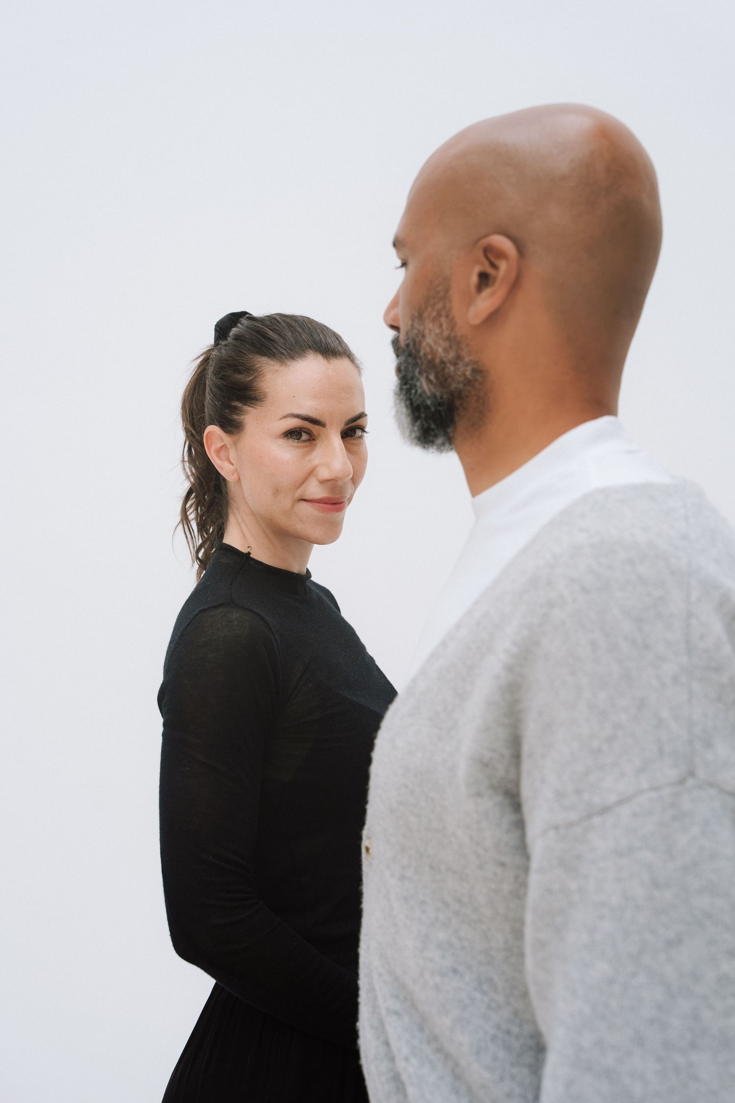 A woman in black and a bald bearded man in gray face each other against a white background.