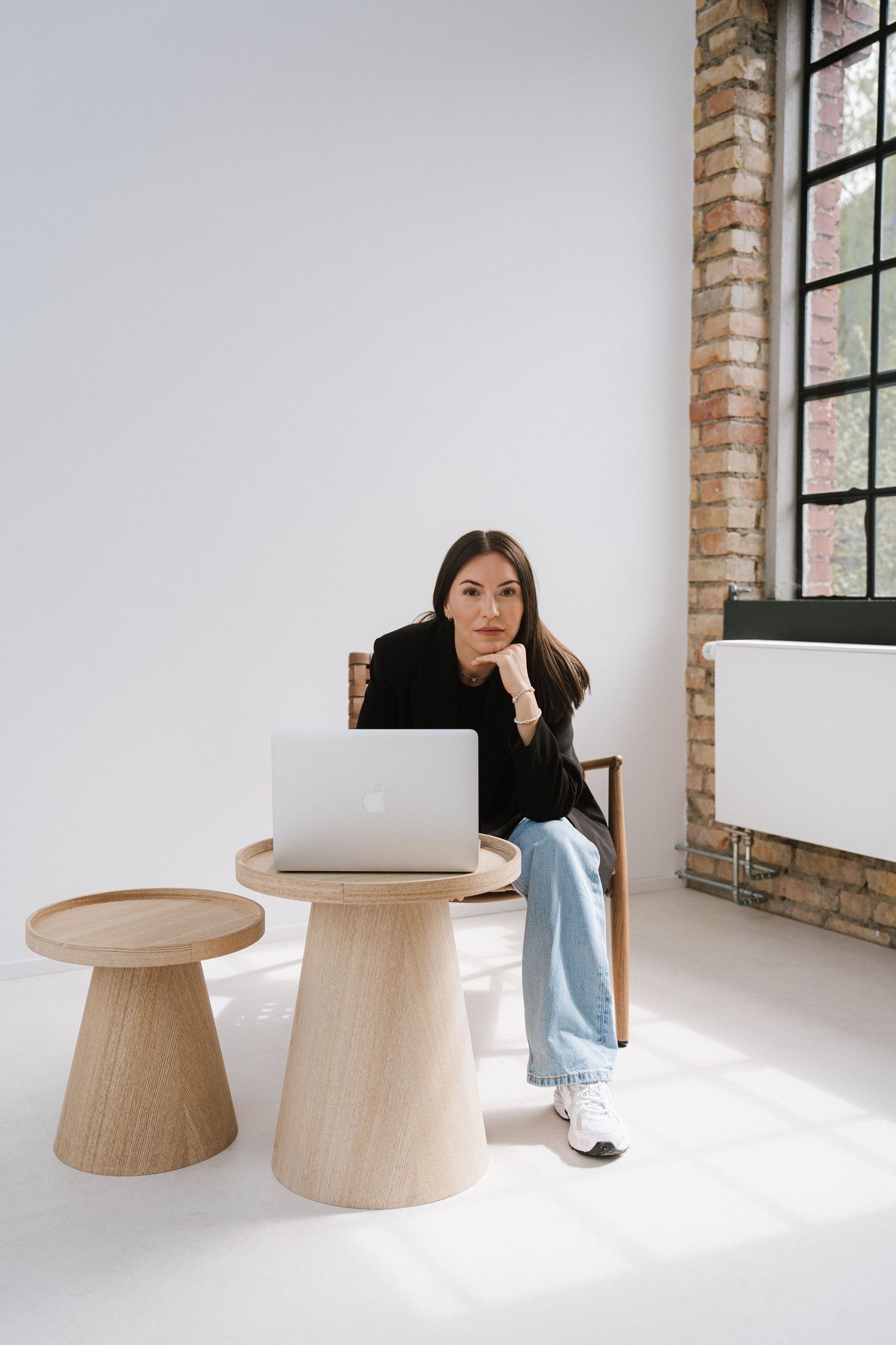 Woman in black blazer and jeans sits at wooden table with laptop in bright modern studio space.