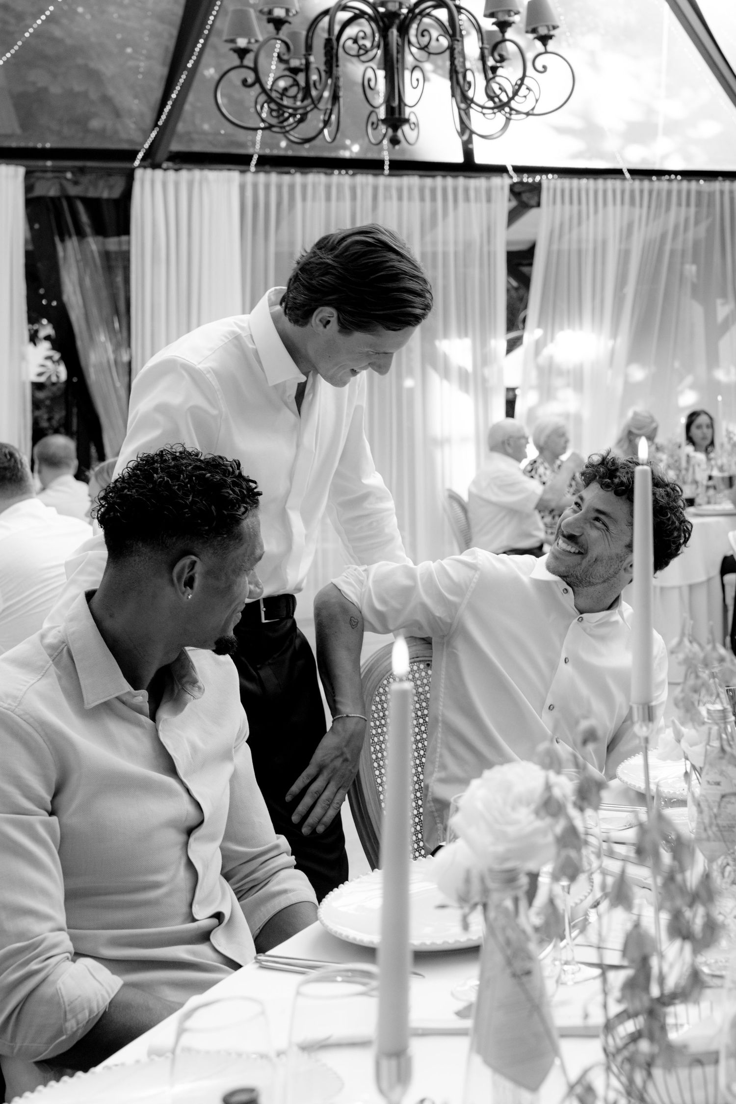 Black and white photo of three men in white shirts sharing a joyful moment at an elegant wedding reception dinner.