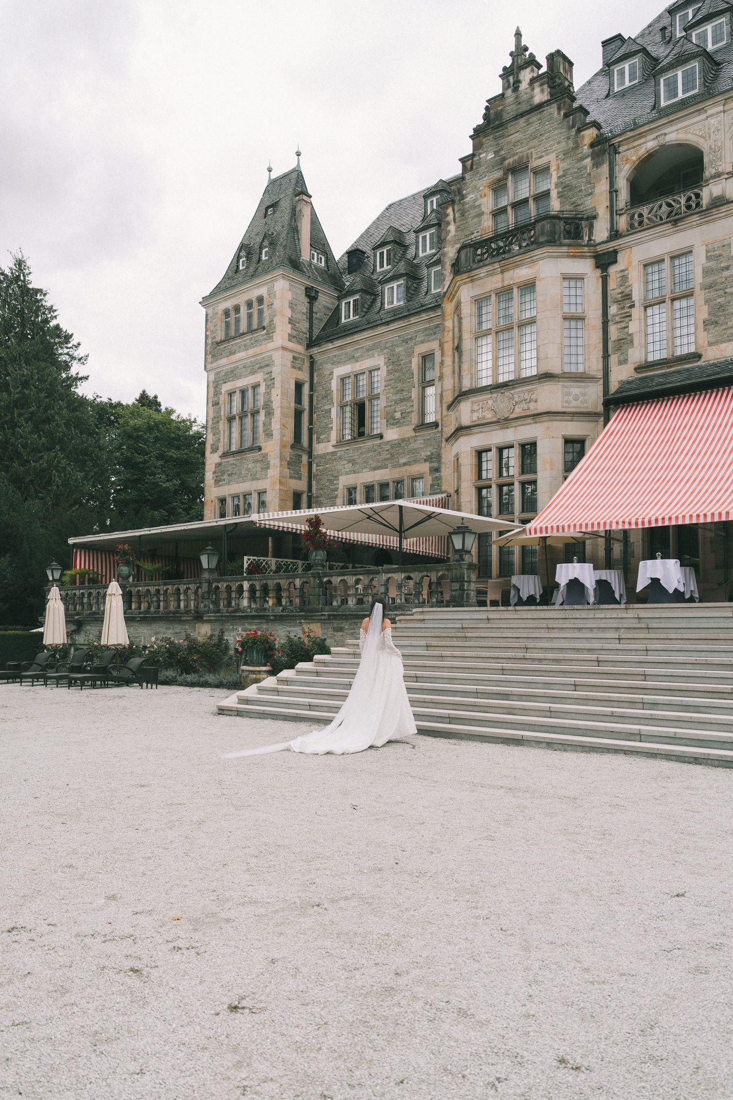 Bride in white gown stands before grand stone castle venue with red awnings and stone steps.