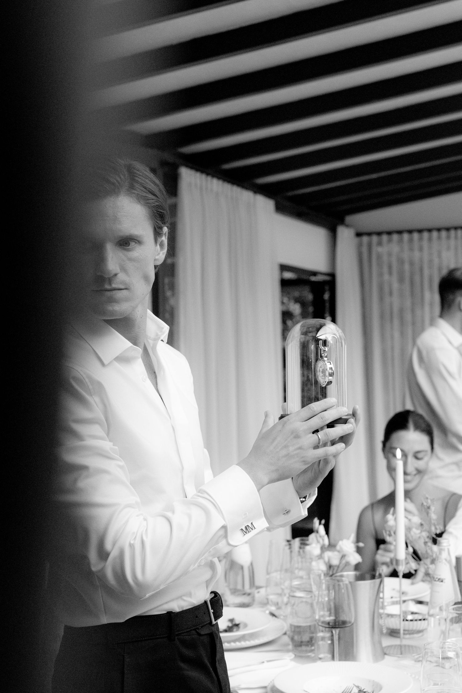 Black and white photo of an elegant man in white shirt holding a bottle at a formal dinner event.