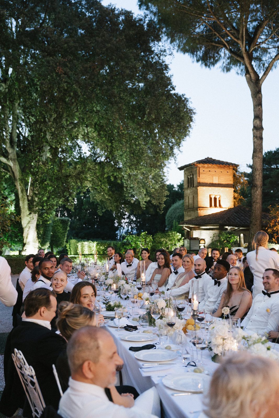 Elegant outdoor wedding reception dinner with guests in formal attire seated at long decorated table in garden setting.