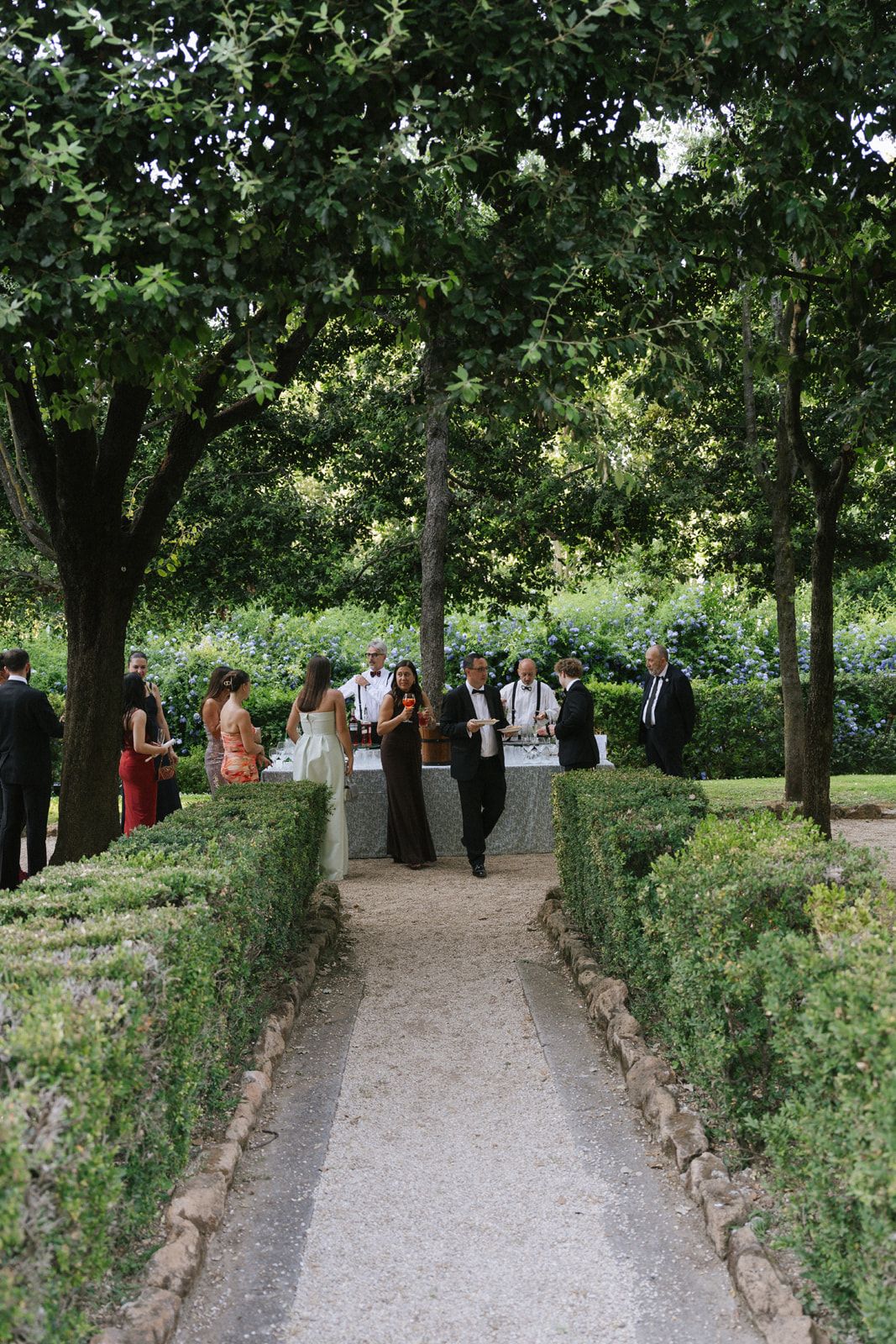 Elegant outdoor garden wedding reception with guests mingling along a tree-lined stone pathway surrounded by manicured hedges.