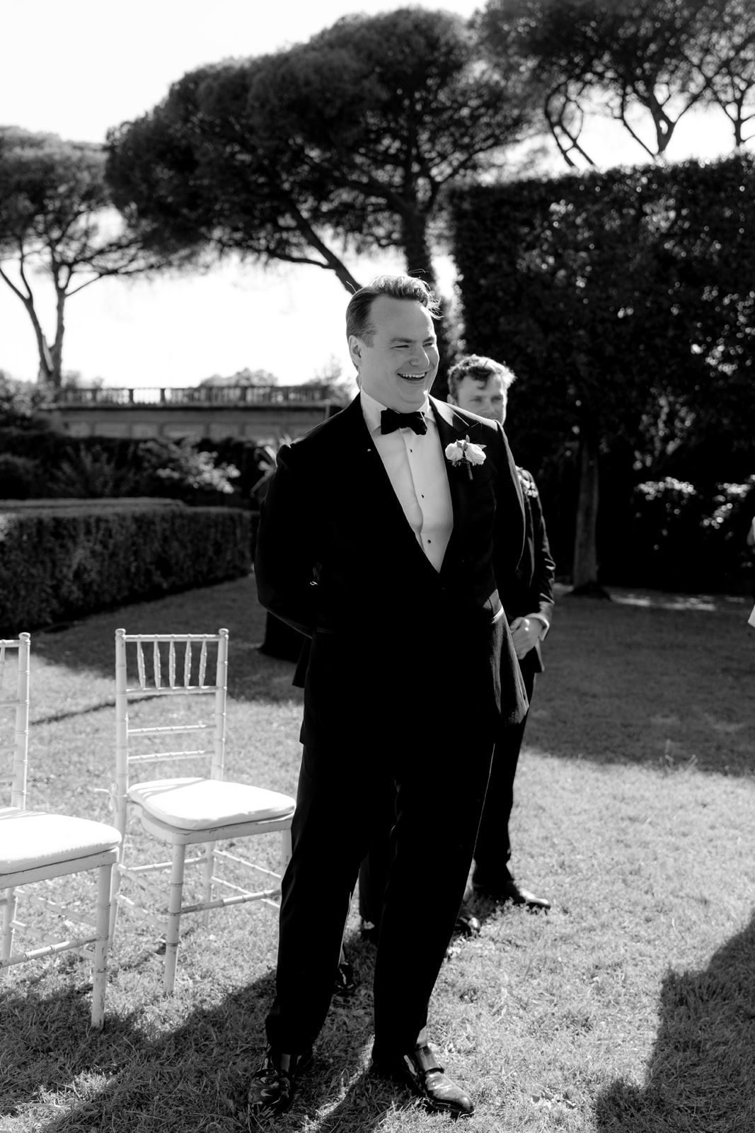 Groom in black tuxedo smiling at outdoor wedding ceremony with manicured garden backdrop, black and white photo.