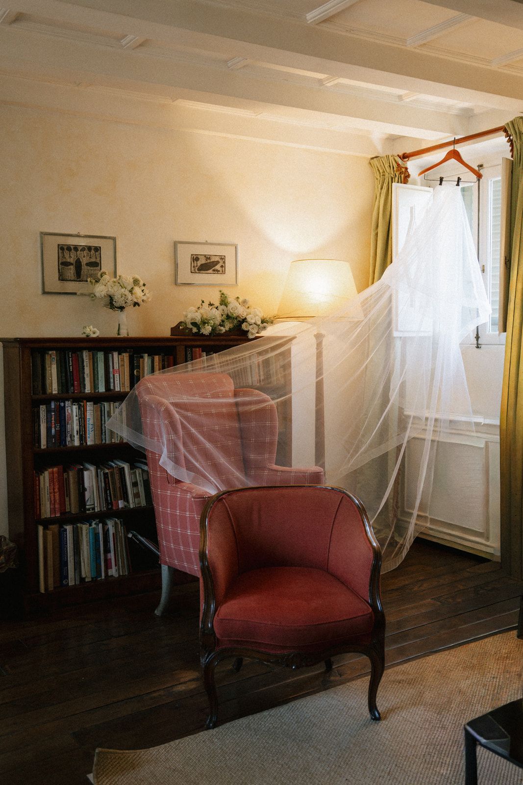 Elegant bridal suite with wedding dress hanging by window, red armchair, bookshelf, and floral arrangements in warm lighting.