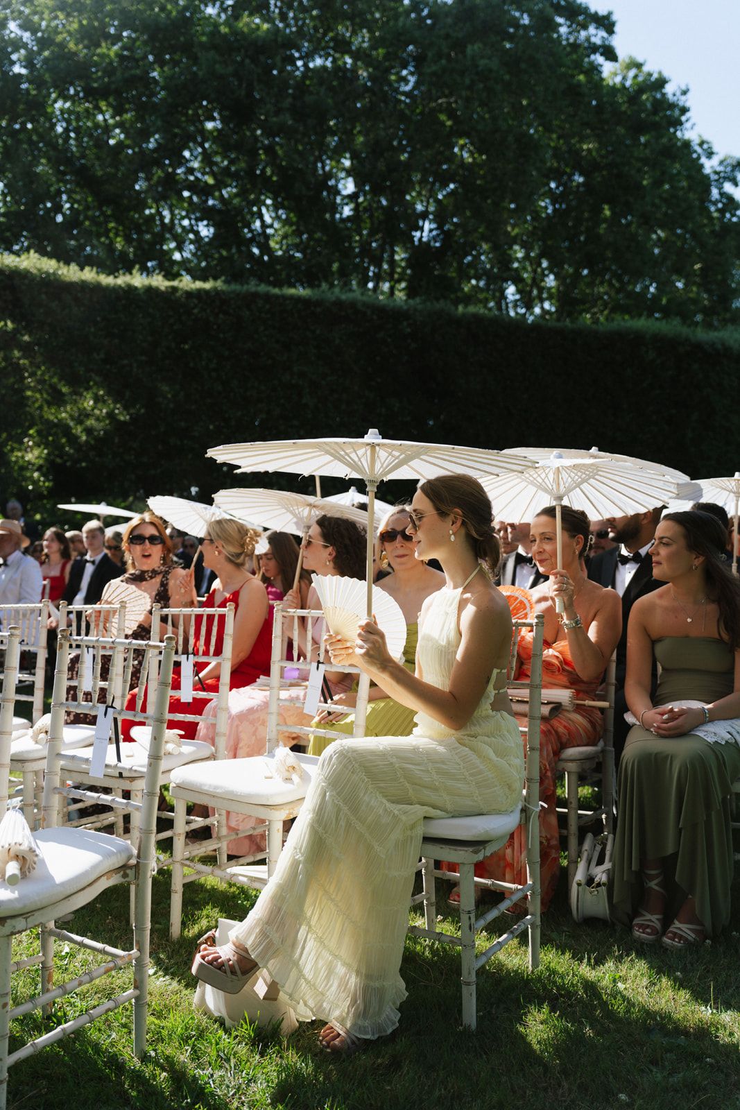 Guests seated outdoors at a garden wedding ceremony, holding white parasols on a sunny day.
