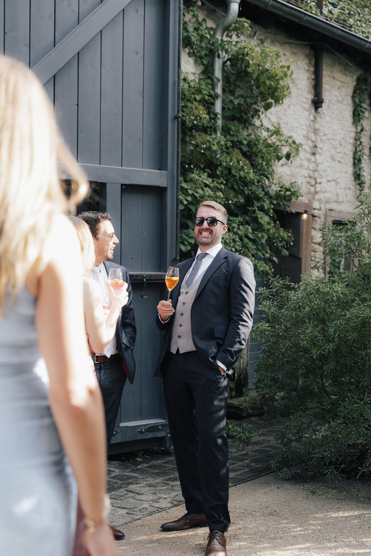 A couple in formal attire stands near a rustic wooden door and stone wall during an outdoor event.