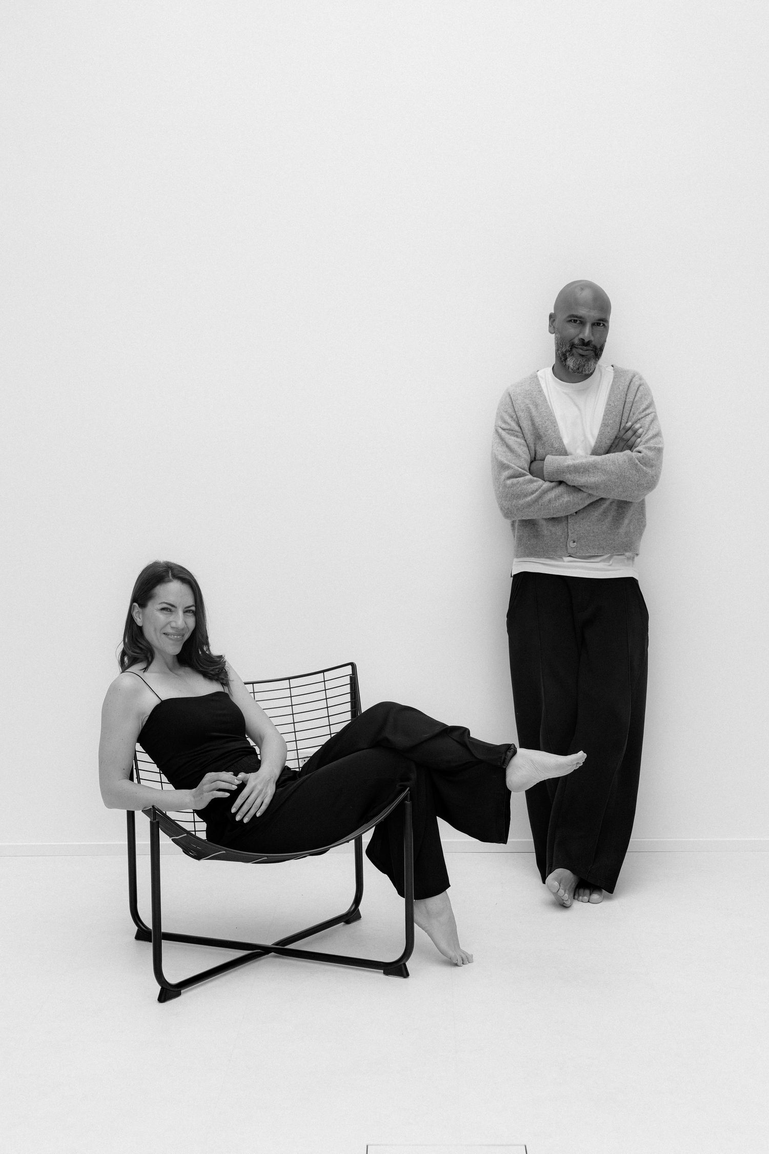 Black and white photo of a woman sitting in a wire chair and a bald man standing with arms crossed against a white wall.