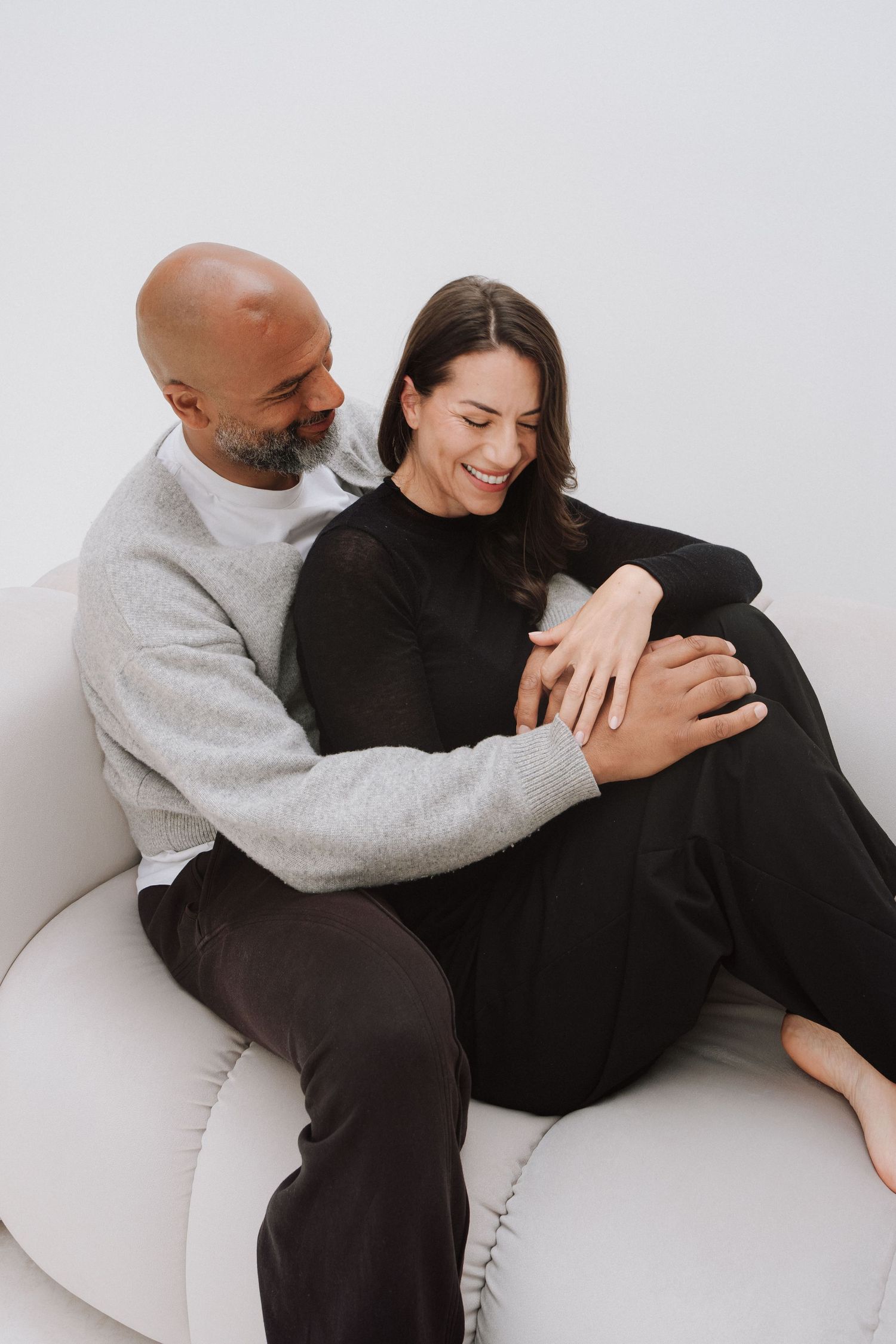 A happy couple sits on a white sofa, embracing warmly, with the woman smiling joyfully in a cozy indoor setting.