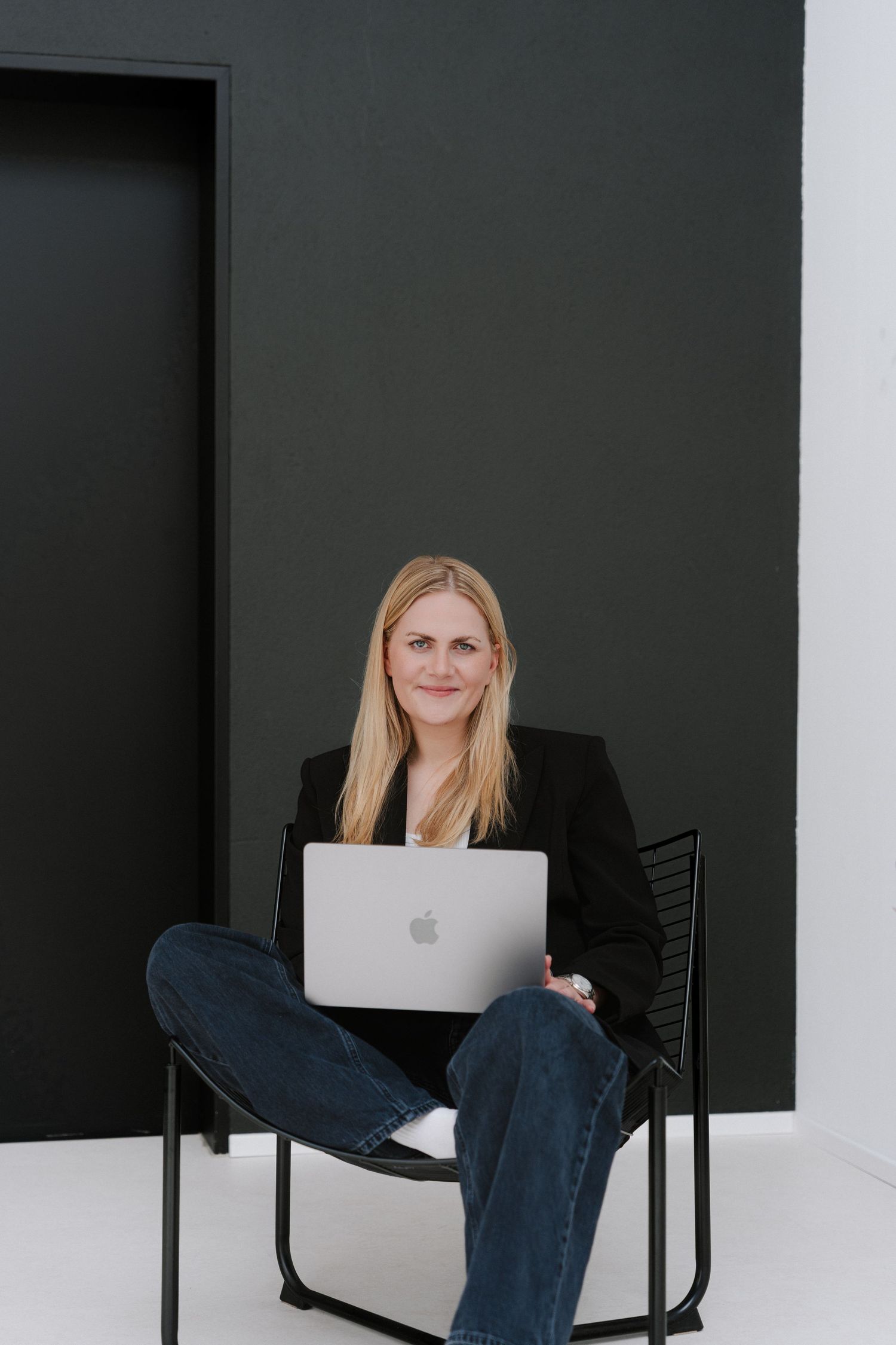 Blonde woman in black blazer and jeans sitting cross-legged on chair with MacBook laptop against dark wall.