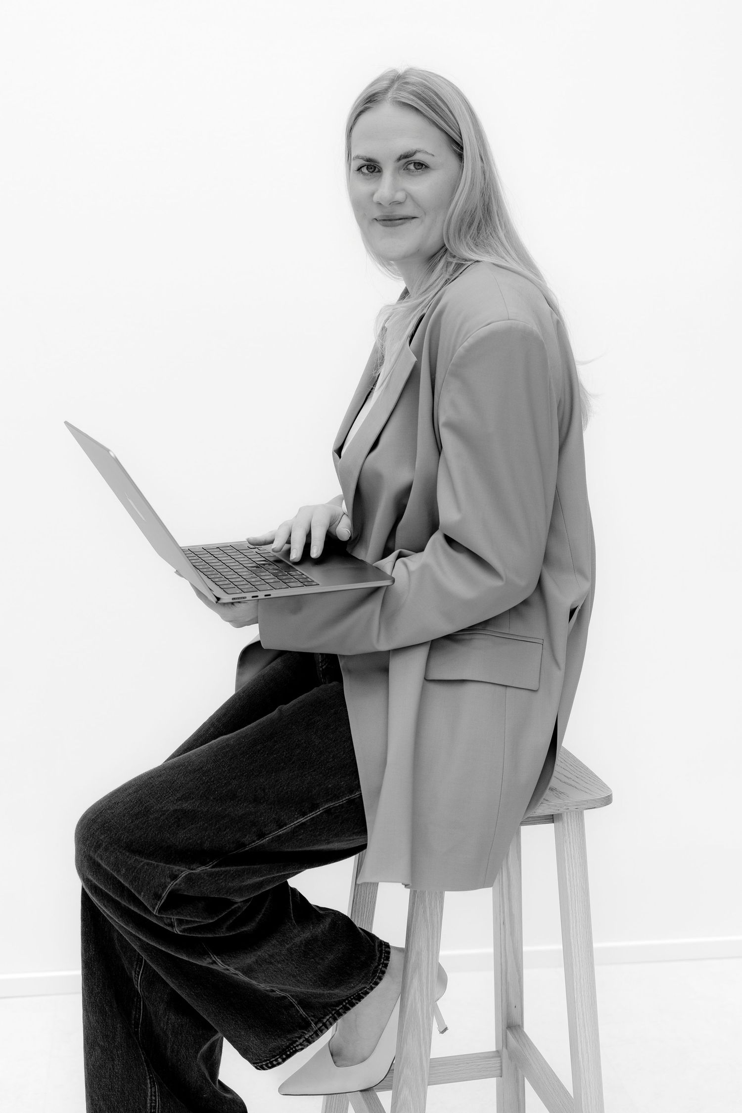 Professional woman in blazer and jeans sits on stool with laptop, black and white portrait photo.