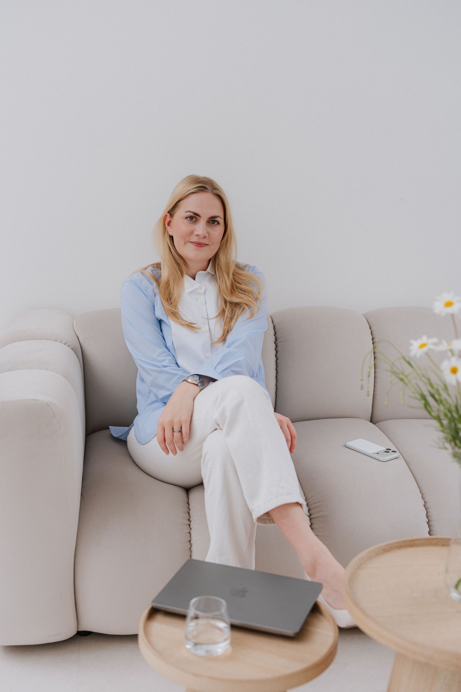 Woman in blue shirt and white pants sitting relaxed on a beige sofa with a laptop on a wooden table.