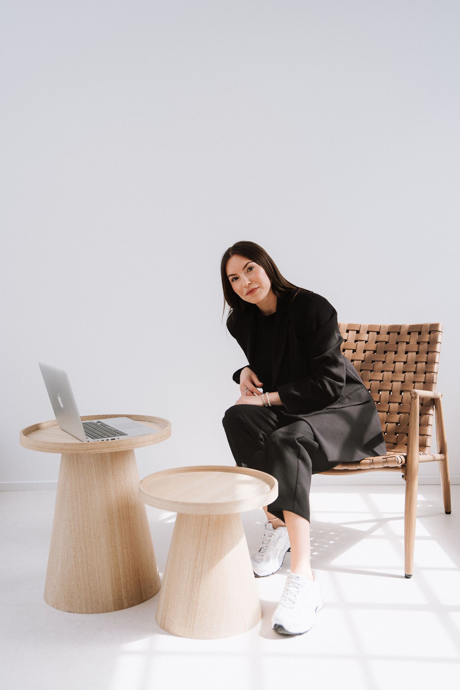 Woman in black outfit sitting on woven chair beside wooden tables with laptop in minimalist white room.