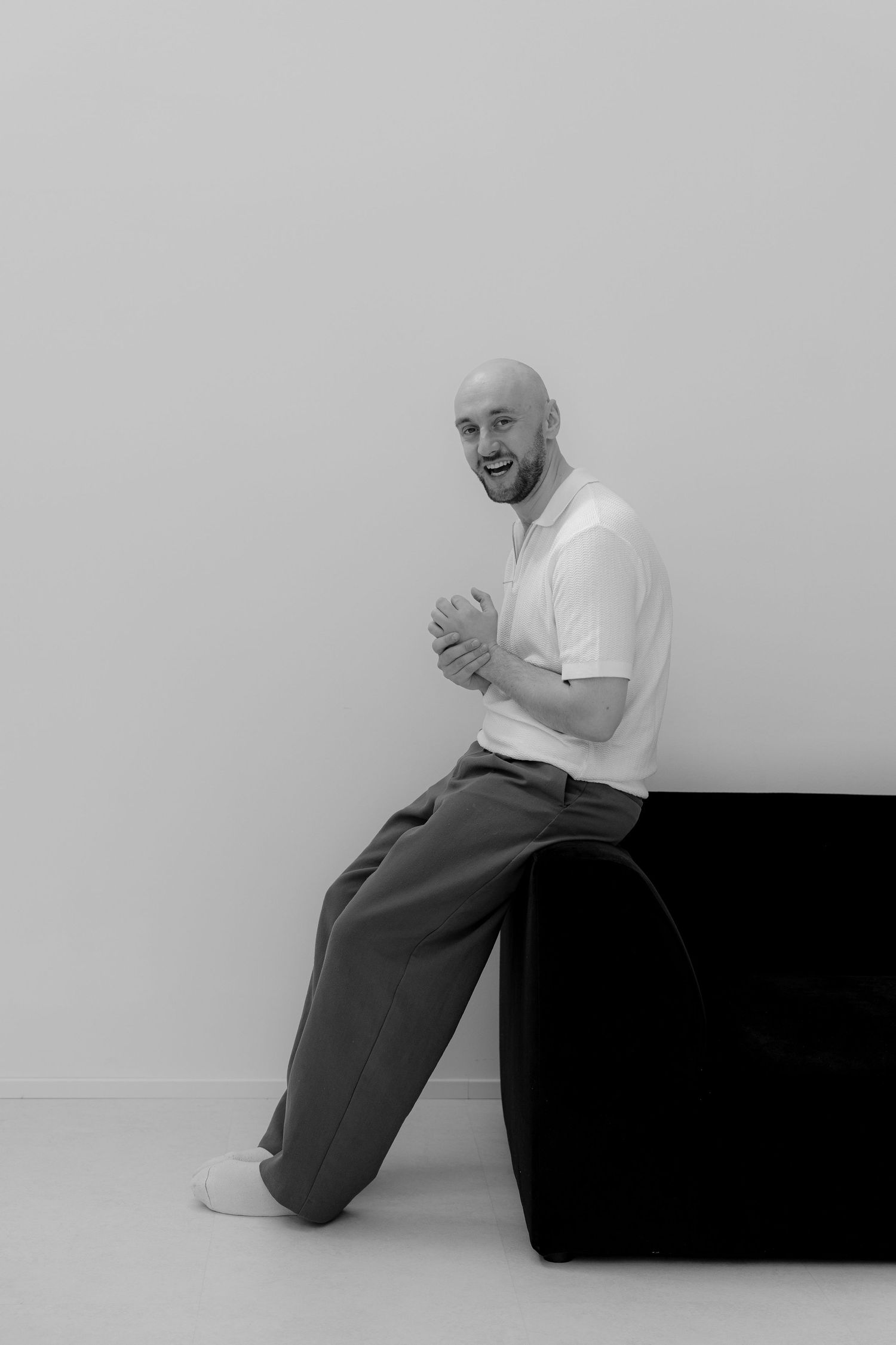 Bald man in white polo and gray trousers smiling while leaning against a black chair in a minimalist studio.