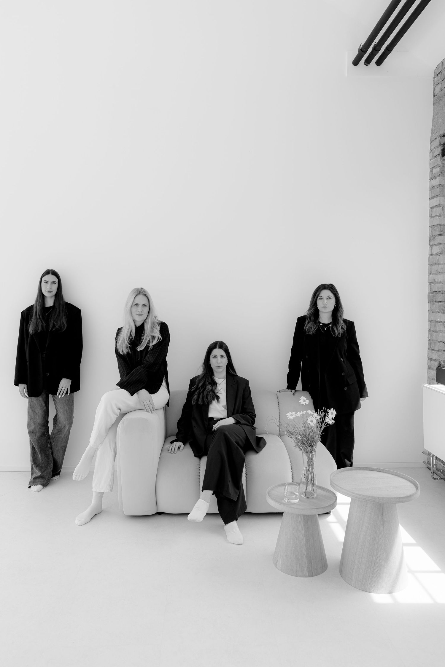Black and white photo of four women in dark blazers posing in a modern minimalist interior with furniture.