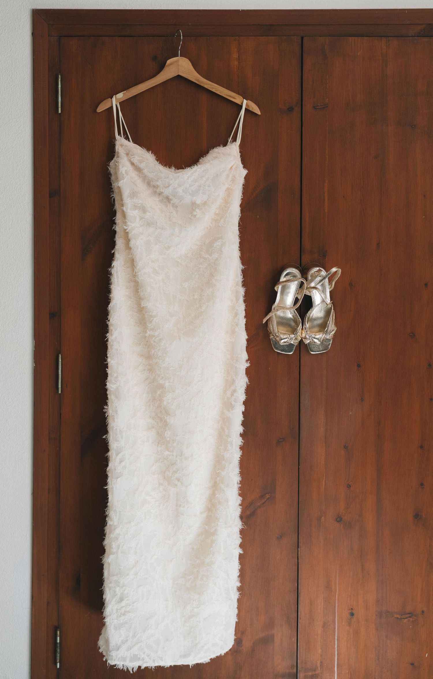 Elegant white wedding dress hanging on wooden door with silver heels.