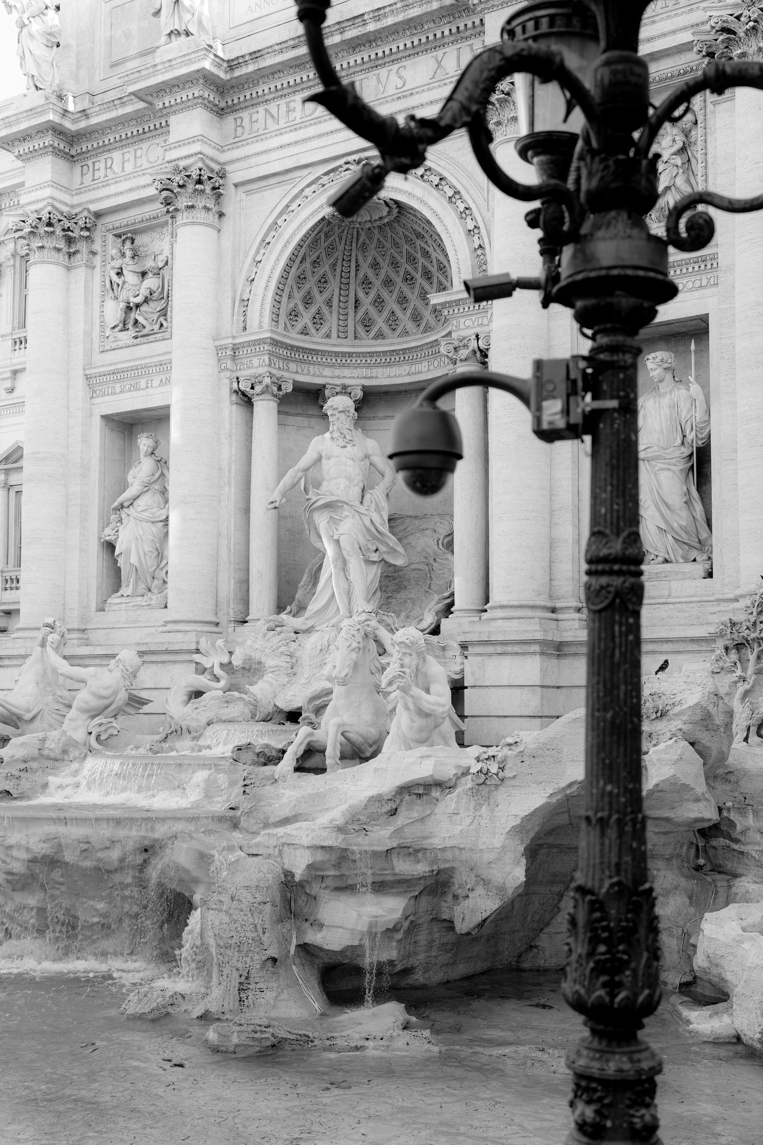 Black and white photo of the Trevi Fountain in Rome, featuring Neptune statue, ornate baroque architecture, and a street lamp.
