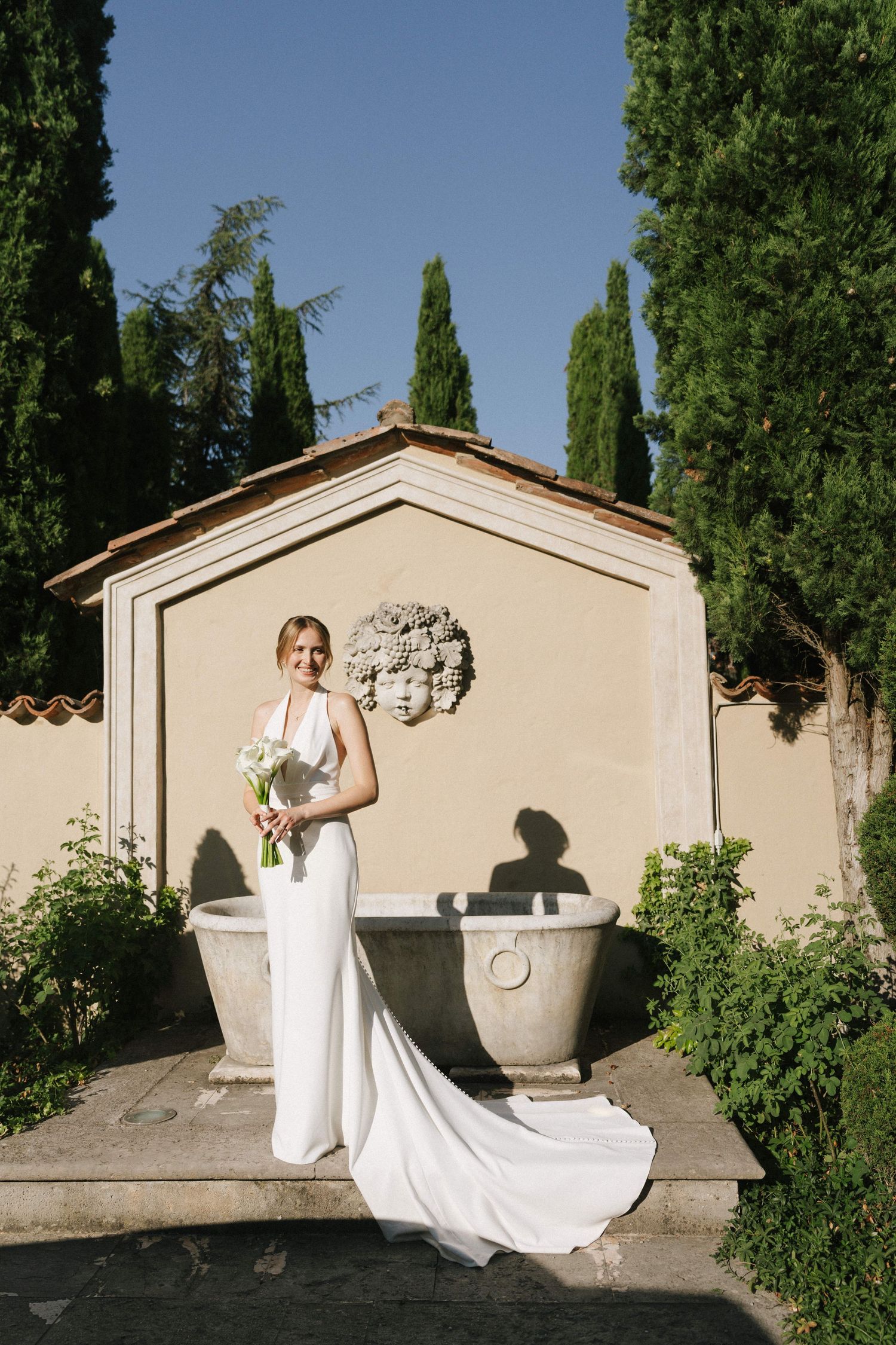 Bride in white gown with train holds bouquet before stone fountain and cypress trees at Italian villa.