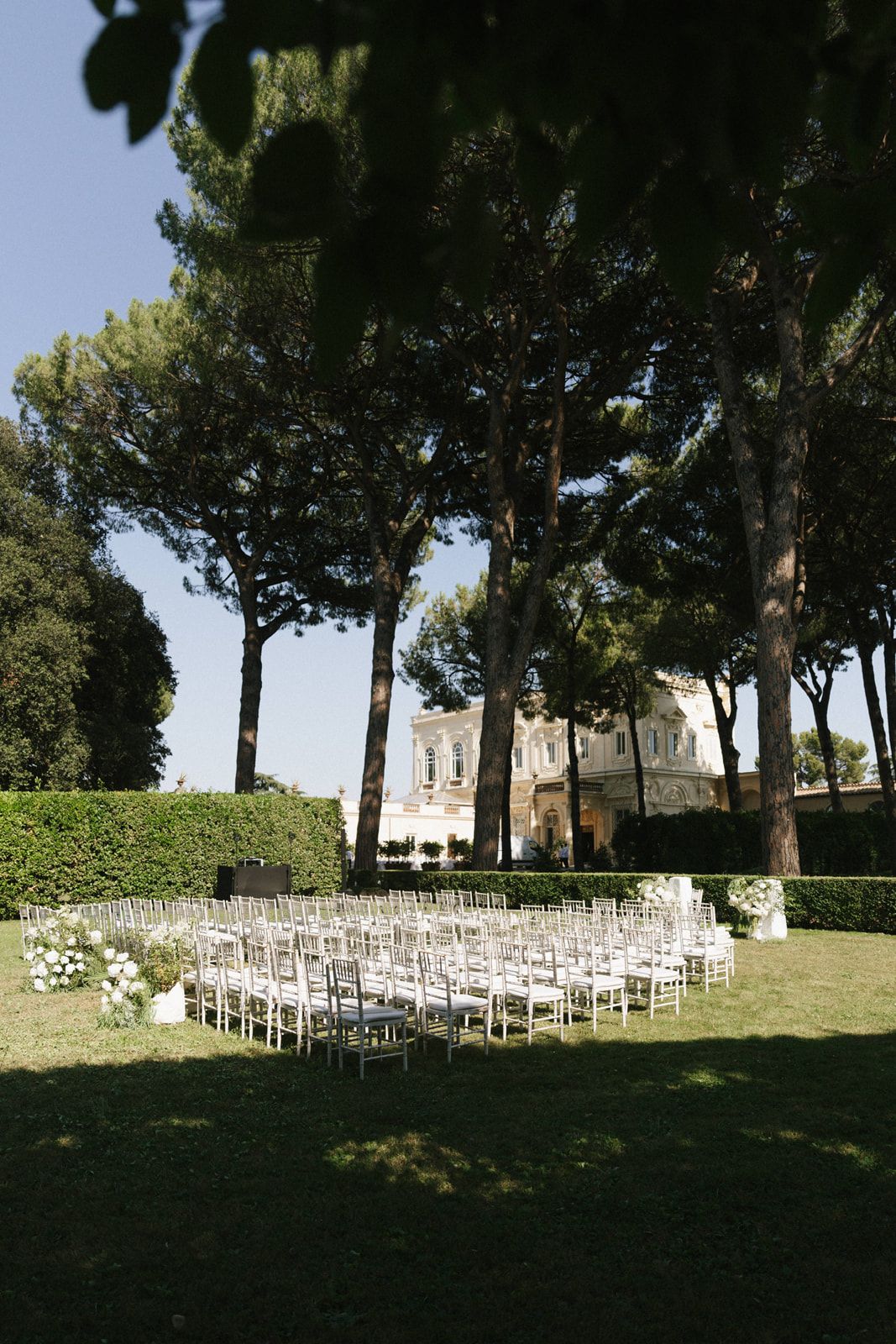 Elegant outdoor wedding ceremony setup with silver chairs on green lawn, tall pine trees, and a historic villa in Rome.