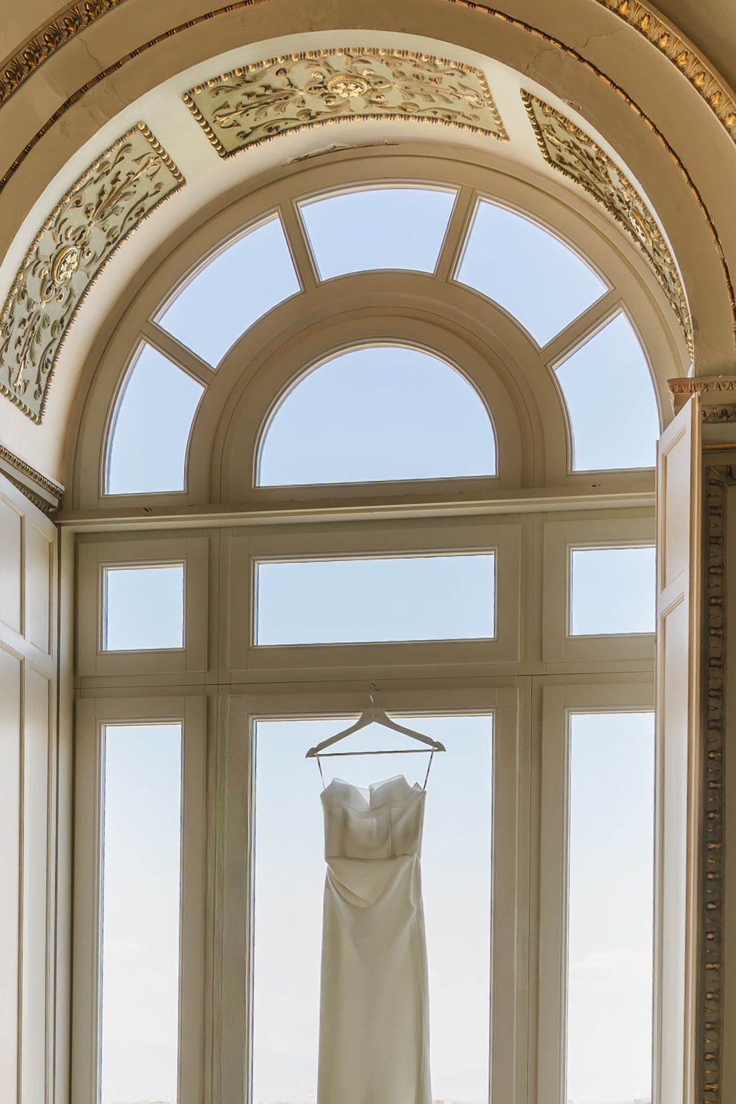 Elegant white wedding dress hanging on a hanger in front of an ornate arched window in a grand venue.
